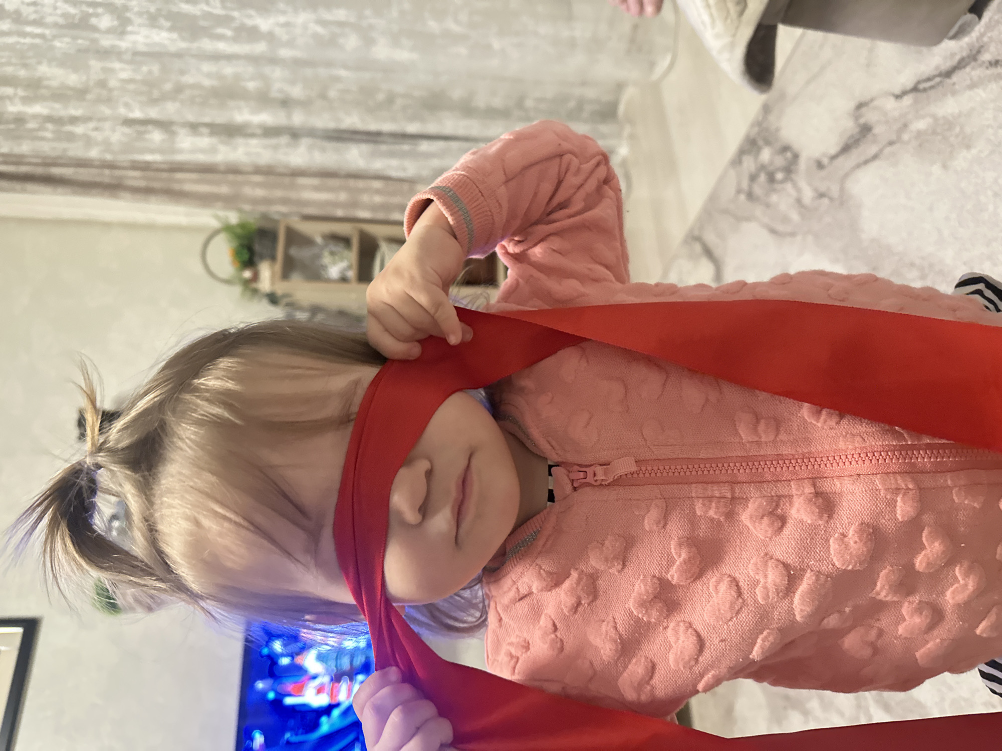 A young child stands indoors, blindfolded with a red cloth, wearing a pink heart-patterned sweater. The location is potentially in Uva, Russia. The background includes a patterned wall, shelves with plants, a television screen displaying blue light, and a light-colored floor. The child is holding the red cloth with both hands.