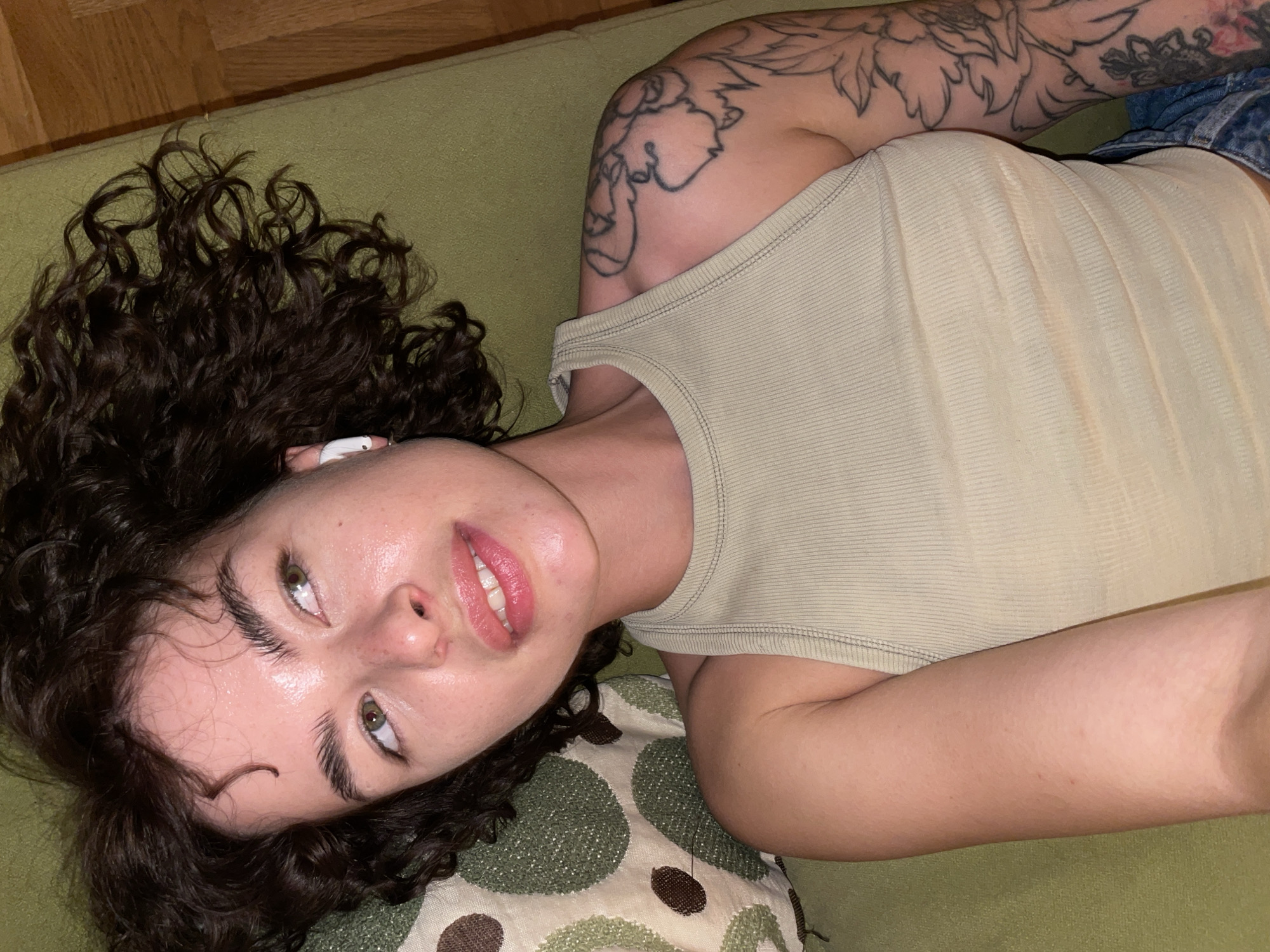 A person is depicted in an indoor setting, reclining on a green, textured surface, likely a sofa or bed, with a wooden floor visible in the background. The individual has dark, curly hair, light skin, and striking green eyes. They are wearing a light beige or pale green ribbed tank top, with a glimpse of denim fabric (possibly jeans) visible towards the lower right. A prominent tattoo featuring floral or leafy outlines covers their left upper arm and shoulder. A white earbud is noticeable in their right ear.
The person is looking slightly upwards and to the side, with a relaxed and possibly playful expression, their lips slightly parted to reveal teeth. Their eyebrows are dark and well-defined, and their skin appears dewy. A patterned cushion with green and brown circular designs is visible beneath their head/shoulder area. The overall mood is casual and relaxed. There is no visible text in the image.
The location context provided is Boreti, Montenegro. The time of day is not explicitly indicated but appears to be well-lit indoors.