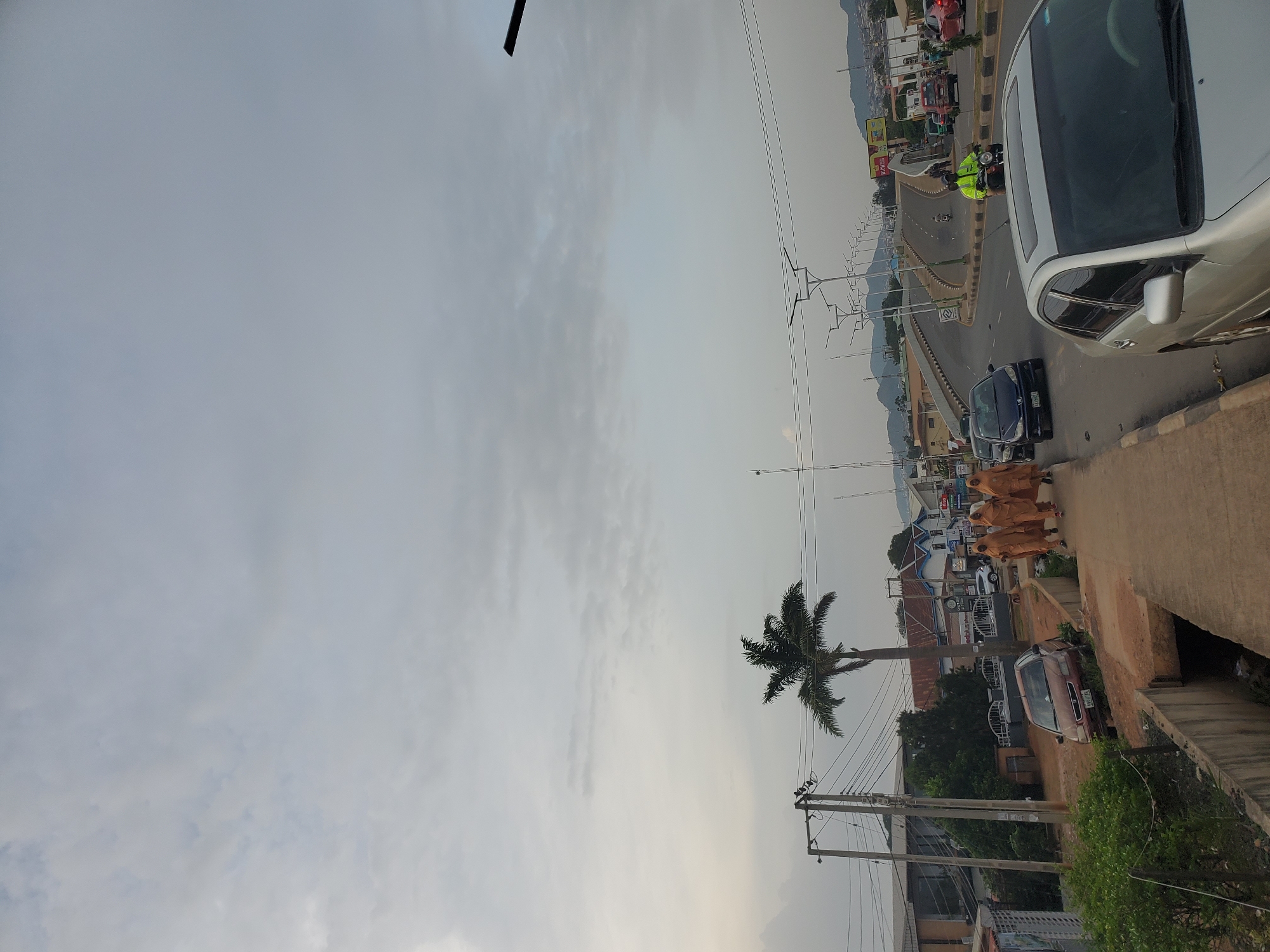 The image depicts an overcast sky over a street scene in Jos, Nigeria. On the left side of the image, there is a paved sidewalk with a concrete drainage channel running alongside it. A red sedan is parked on the dirt shoulder of the road, with a dark sedan parked behind it. Three women, wearing light brown hijabs and dresses, walk on the sidewalk towards the right. A palm tree stands behind them, with telephone poles and wires in the background. The road in front of them curves to the right, with several cars, including a silver sedan in the foreground, and a motorcycle with a rider wearing a bright green jacket. In the distance, there are buildings and hills, with streetlights lining the road. A billboard with text is visible on the right side of the road.