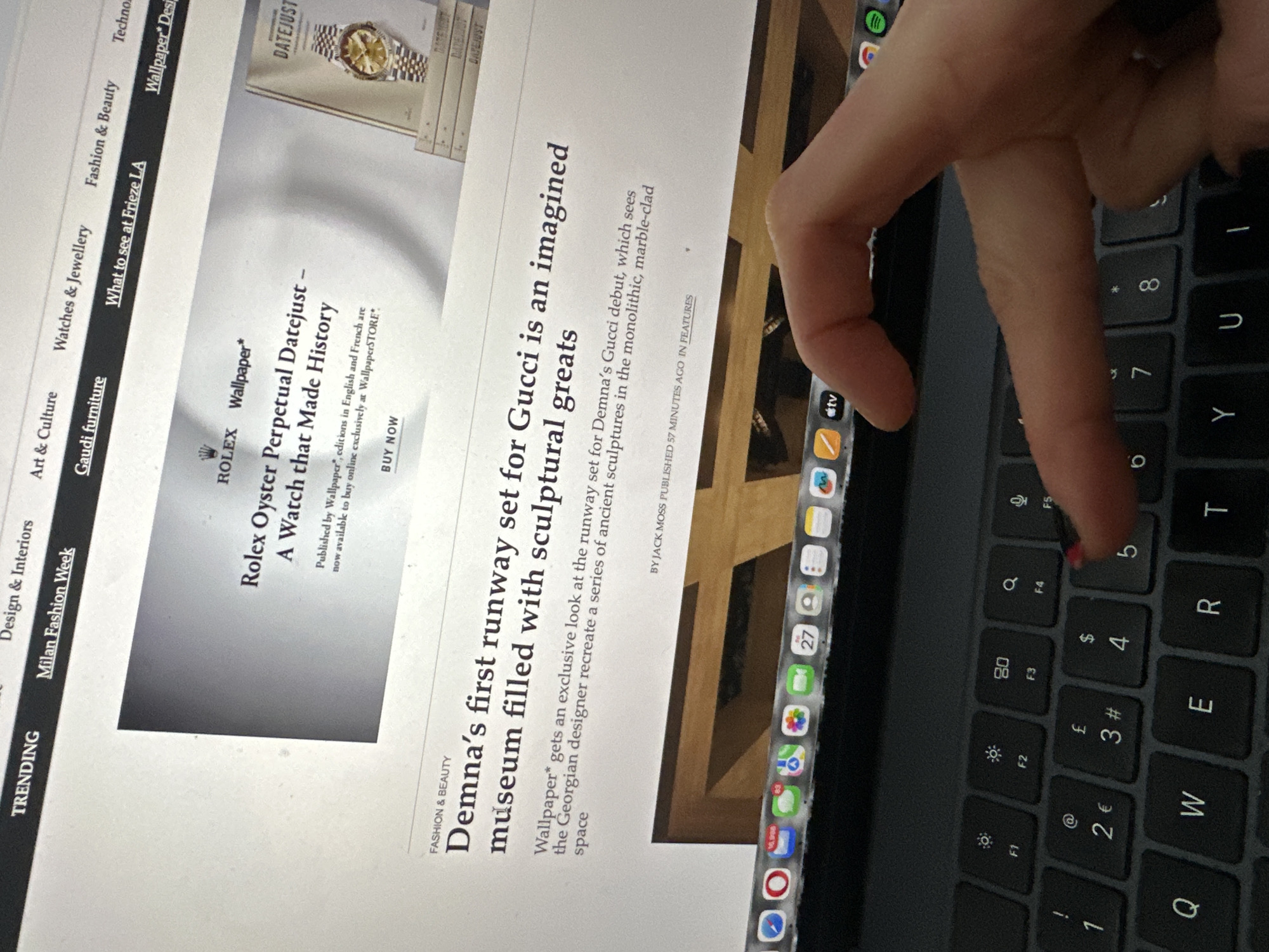 The image displays a close-up, angled view of a human left hand, with red nail polish on the index finger, poised over the '5' key of a black QWERTY laptop keyboard. The laptop screen shows a "Wallpaper*" website featuring an advertisement for a "Rolex Oyster Perpetual Datejust" watch and an article titled "Demna's first runway set for Gucci is an imagined museum filled with sculptural greats." The website's navigation bar is visible at the top, showing categories such as "Trending," "Milan Fashion Week," "Design & Interiors," "Art & Culture," "Watches & Jewellery," "Fashion & Beauty," and "What to see at Frieze LA." The laptop's application dock is partially visible at the bottom of the screen, displaying icons including Safari, Mail (with numerous unread messages), Messages, Calendar, and TV. The scene is located in the city of Wealden, United Kingdom.