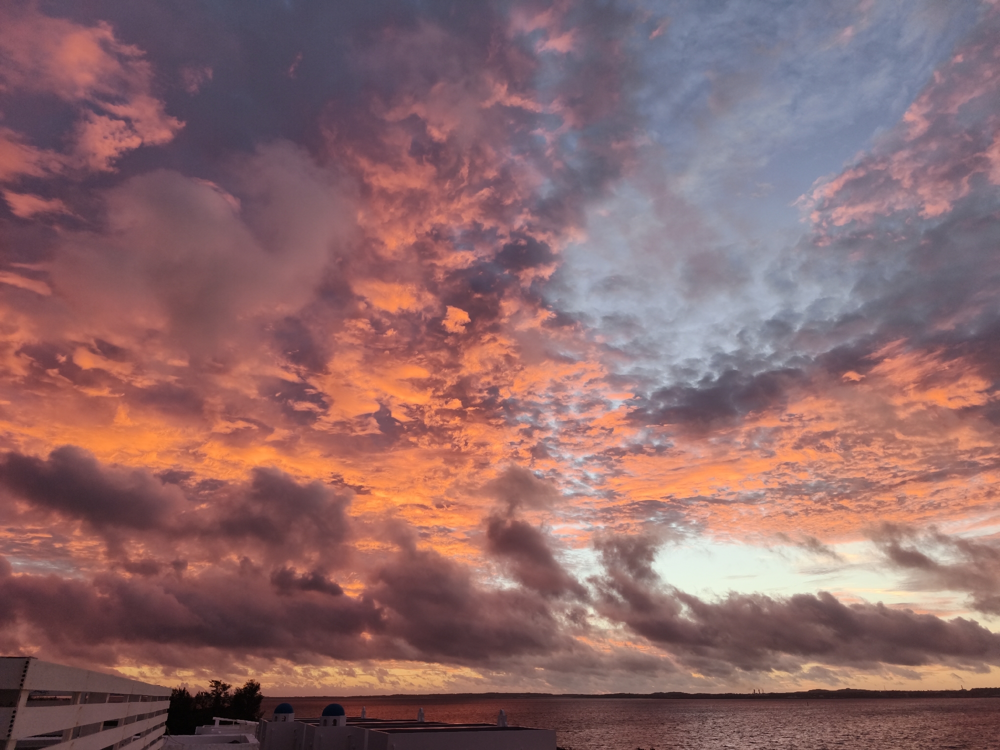 The image captures a dramatic sunset over the ocean in Miyakojima, Japan. The sky is filled with vibrant hues of orange, pink, and purple, illuminated by the setting sun. Fluffy clouds are scattered across the sky, creating a dynamic and textured canvas. In the foreground, a white railing runs along the edge of a building, with hints of blue-domed structures visible. The ocean reflects the colors of the sky, creating a serene and beautiful atmosphere. The distant horizon is lined with silhouetted trees and landforms, adding depth to the scene.
