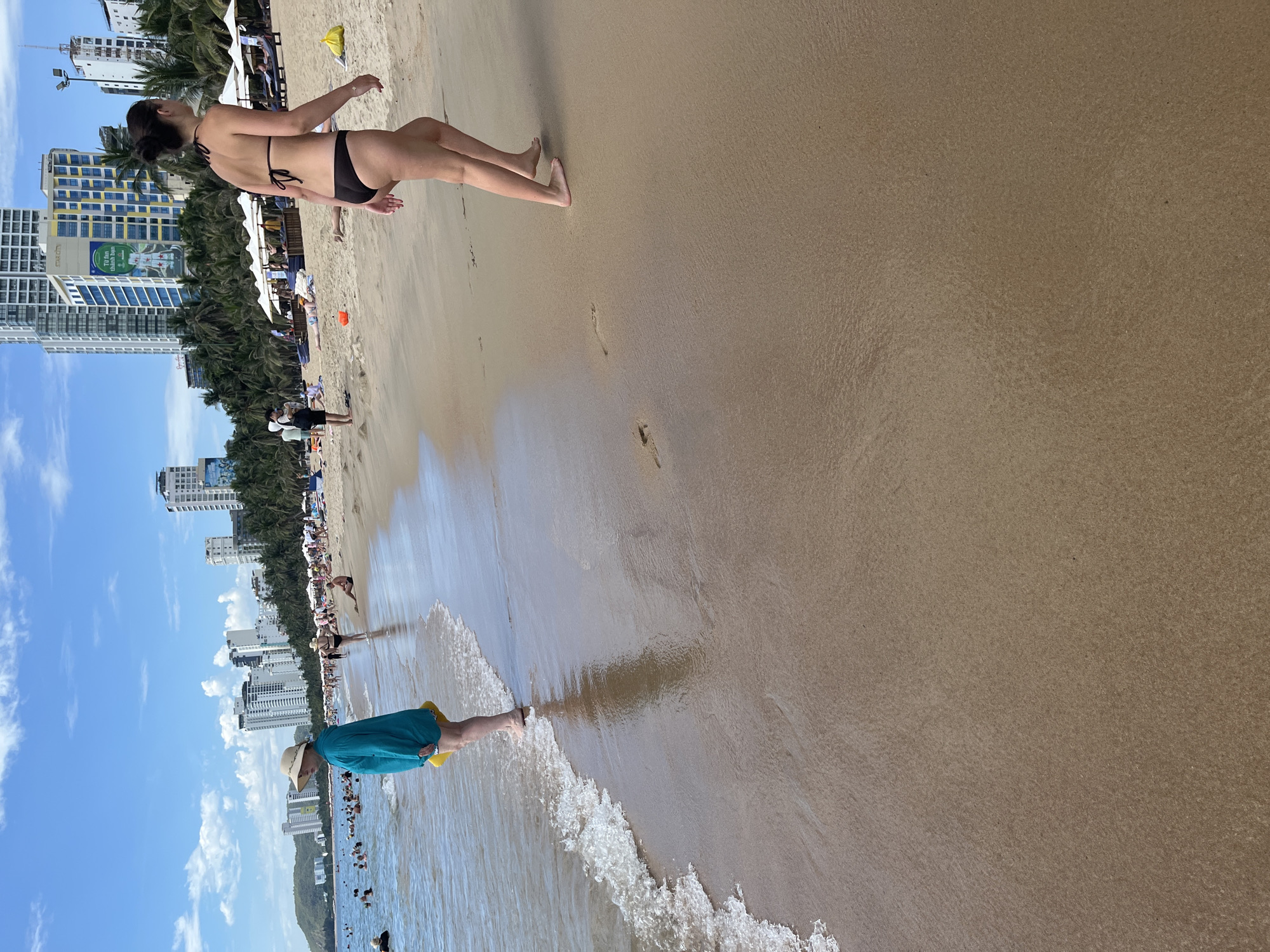 The image captures a beach scene in Nha Trang, Vietnam, likely during daylight hours given the clear blue sky with scattered clouds. The foreground is dominated by the wet sand of the beach, with gentle waves lapping at the shore.
In the mid-ground, a person wearing a wide-brimmed straw hat and a teal long-sleeved garment stands at the edge of the water, holding a yellow object. Further along the beach, a woman in a dark bikini walks away from the camera. She has dark hair pulled back and is barefoot.
In the background, the beach is populated with other beachgoers, some wading in the water, others relaxing on the sand or under umbrellas. Numerous high-rise buildings line the coast, characteristic of Nha Trang's urban beachfront. Palm trees are also visible, suggesting a tropical environment. The overall impression is a busy, sunny day at a popular beach destination.