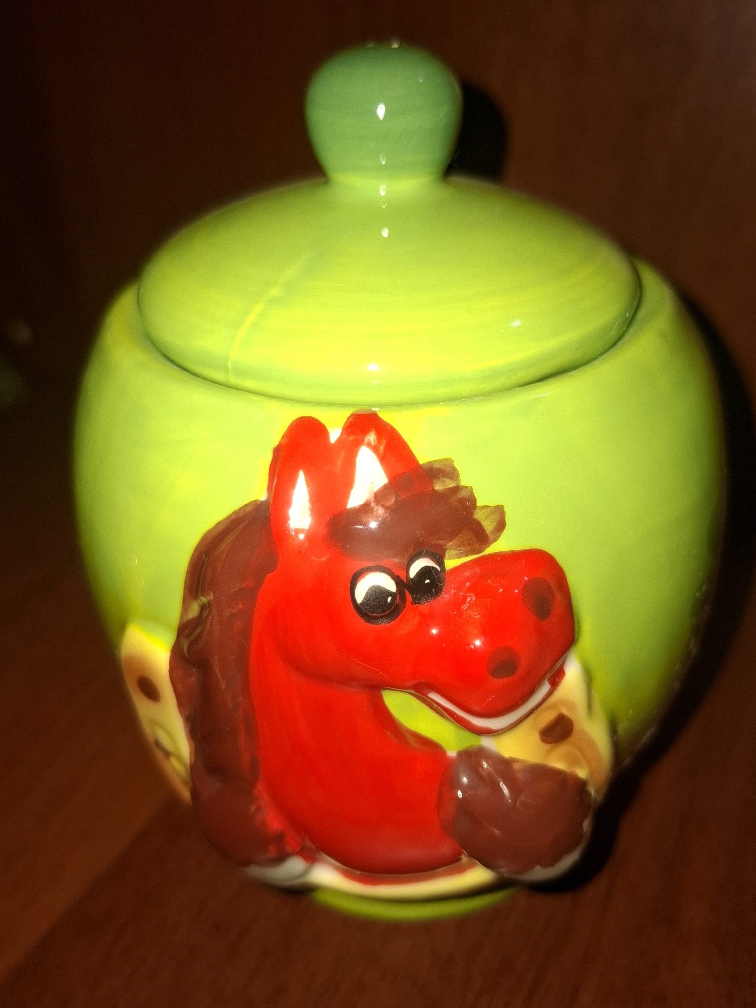 An overhead shot captures a vibrant lime-green ceramic jar with a playful red horse design adorning its front. The jar is topped with a matching green lid, featuring a small, rounded knob. The horse's face is rendered with large, expressive black and white eyes and a red snout dotted with darker red spots. Its mane and ears are depicted in a rich brown. The jar itself rests on a warm, brown wooden surface, likely a table or countertop. The lighting is somewhat dim, casting subtle shadows and highlighting the glossy sheen of the ceramic. The background is a dark, indistinct brown, suggesting an indoor setting. This item does not appear to be located in Almaty, Kazakhstan.