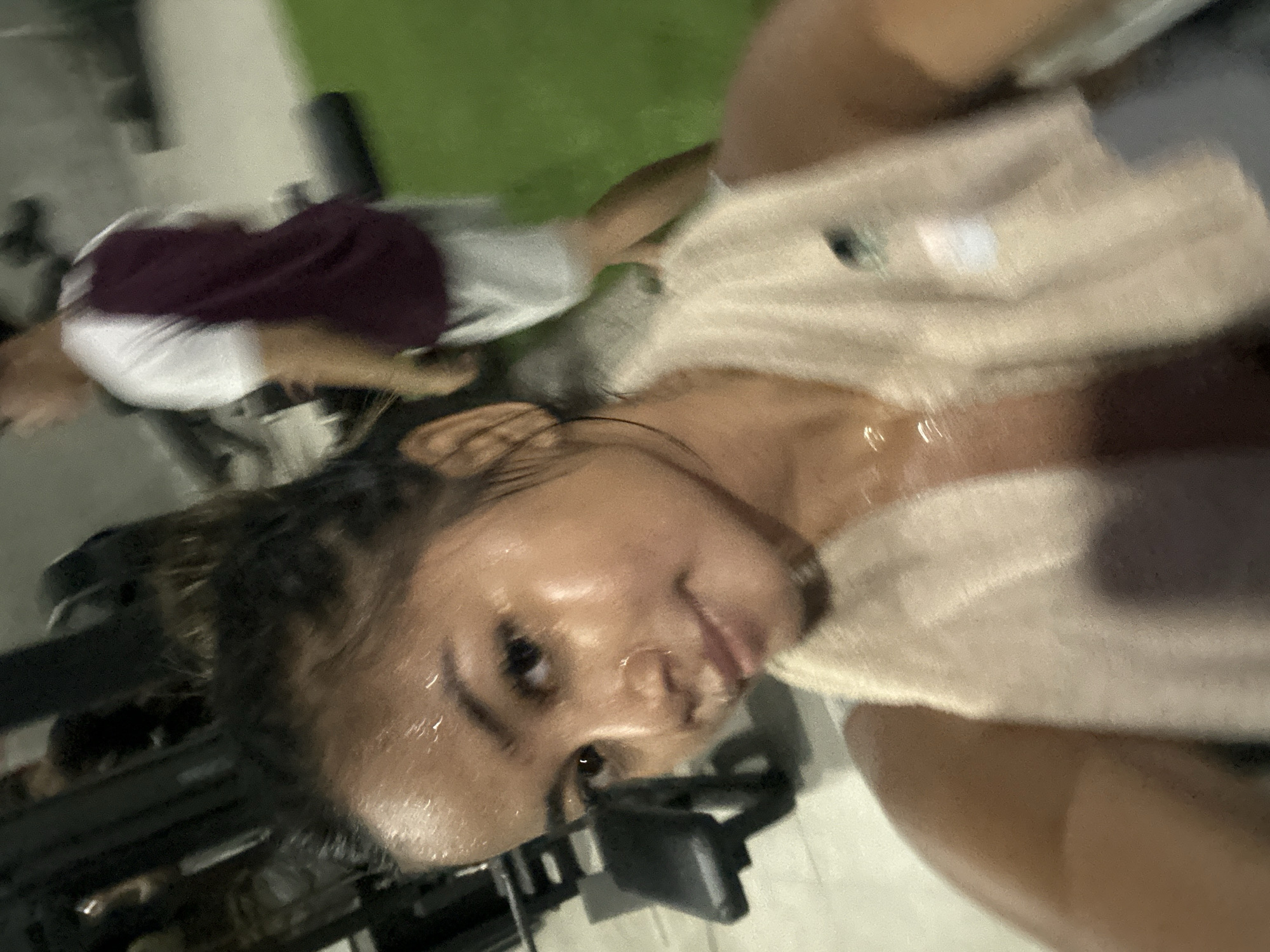 A close-up, slightly blurred selfie shows a woman with a towel draped around her neck, her face and arms glistening with sweat. She is in what appears to be a gym, with weightlifting equipment visible in the background. To her right, a man in a maroon and white shirt and grey shorts is also visible, slightly out of focus. The environment suggests an indoor fitness facility, possibly located in Sukawati, Indonesia, given the metadata. The woman's expression is relaxed, implying she has finished a workout.