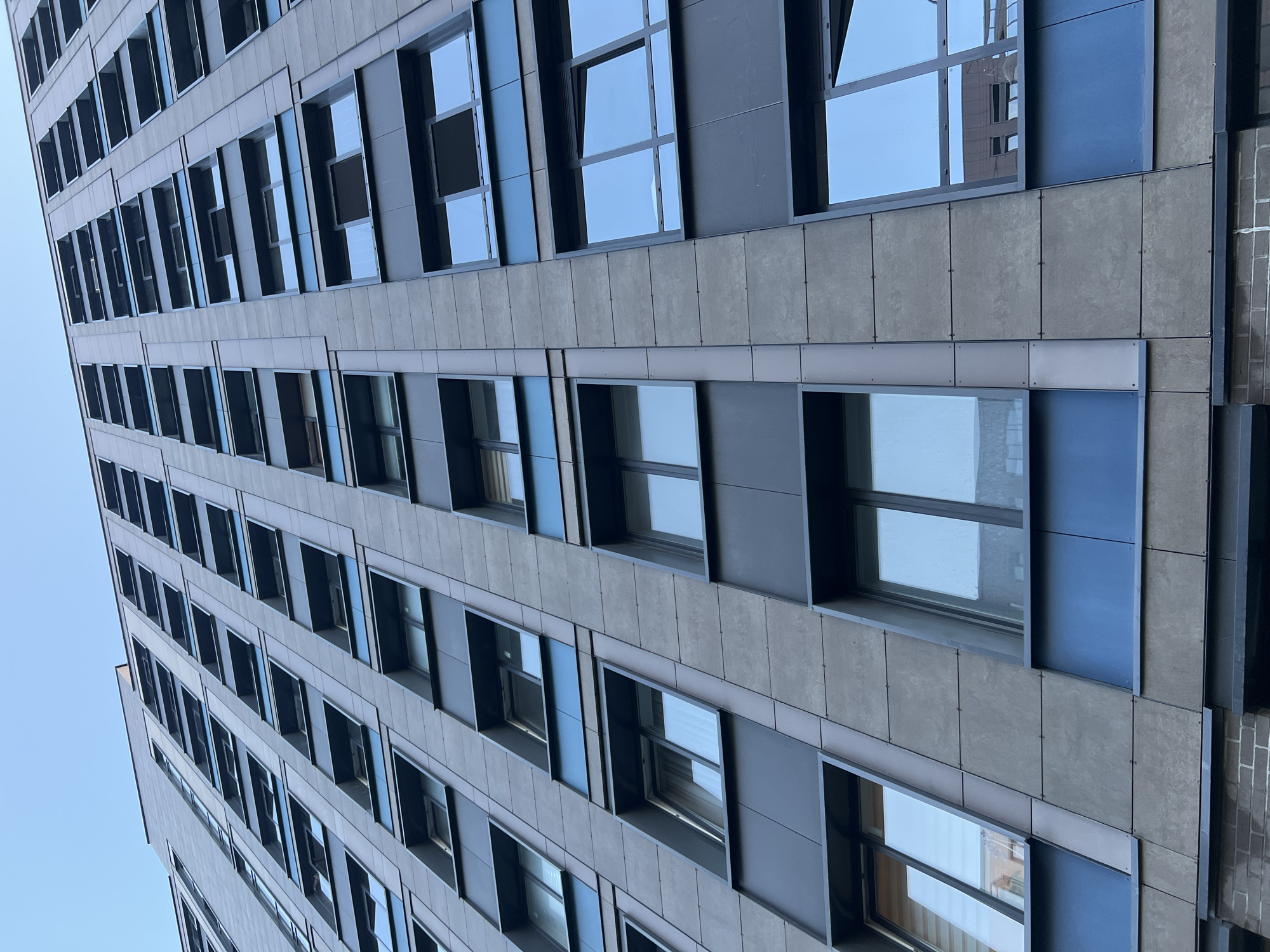 The image shows a close-up, low-angle view of a modern, high-rise building with a facade of grey panels and dark blue accents. The building has many windows, some of which reflect the sky or neighboring buildings. The windows are arranged in a grid pattern, with some recessed and others flush with the facade. The overall impression is one of a contemporary urban environment. The specific location is the Nevsky district in Saint Petersburg, Russia, a vibrant area known for its mix of historic and modern architecture.