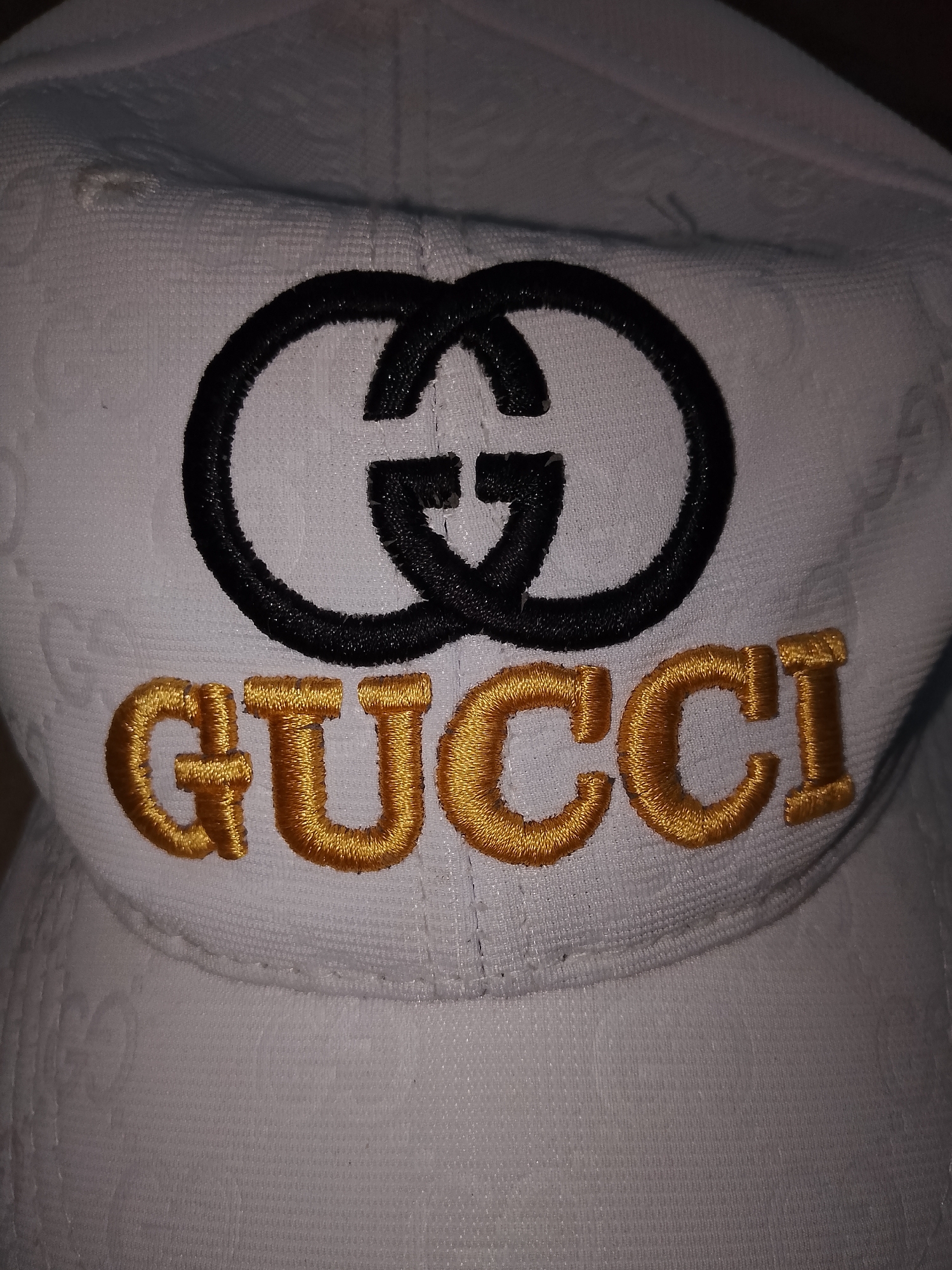 The image displays a close-up, frontal view of a white baseball cap featuring prominent black and gold embroidered branding. The cap's fabric is a textured white material, subtly embossed with a faint, repeating pattern of the interlocking "GG" monogram. Centered on the front of the cap, a large interlocking "GG" logo, representing Guccio Gucci, is densely embroidered in black thread, exhibiting a slight sheen. Directly beneath this black logo, the word "GUCCI" is embroidered in a rich, golden thread, with each letter densely stitched and appearing raised from the cap's surface. The cap's brim is visible at the bottom of the frame, showing a stitched seam where it meets the crown. The lighting is direct, highlighting the texture of the fabric and the embroidery.