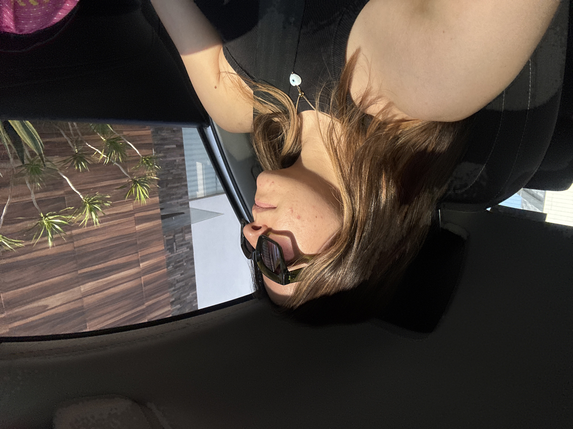 A close-up photograph, correctly oriented, depicts a young person, likely female, from the chest up, leaning back within the dark interior of a vehicle. Her head is tilted to her left, and she appears to be looking away from the camera. She has medium-length, light brown hair and is wearing a dark, possibly black, sleeveless top. A delicate necklace with a light blue and white evil eye-shaped pendant is visible around her neck. Dark-framed sunglasses rest on her nose and cheeks. Bright sunlight illuminates her face and arm, casting strong shadows, indicating a clear, sunny day.
Through the vehicle's window, an outdoor scene is visible. It features a building with a textured wall, resembling brown wood paneling, set against which are several green, spiky-leaved plants. Further back, another part of the building shows a grey, possibly stone or concrete, block facade. The overall environment suggests a modern architectural setting, potentially a garden or patio area, consistent with a warm climate like Puebla, Mexico. The person appears relaxed. No text is visible in the image.