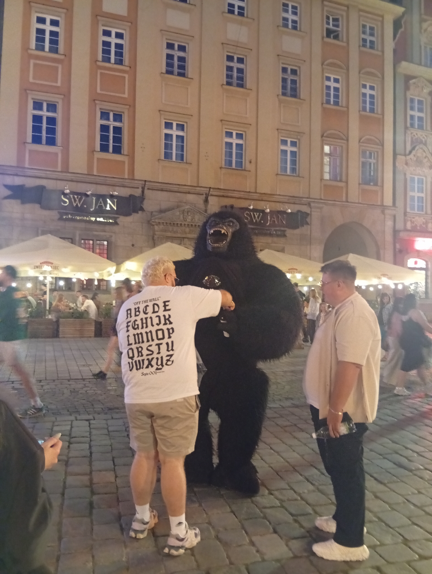 In Wrocław, Poland, on a cobblestone street outside a building with a sign reading "SW. JAN", a person in a gorilla costume is interacting with two men. The man closer to the viewer, wearing a white t-shirt with the alphabet printed on it and beige shorts, stands facing the gorilla. To his right, another man in a light-colored short-sleeved shirt and dark pants faces the gorilla. In the background, several other individuals are visible, some seated at tables under outdoor canopies. The scene appears to be a street performance or a promotional event at night.