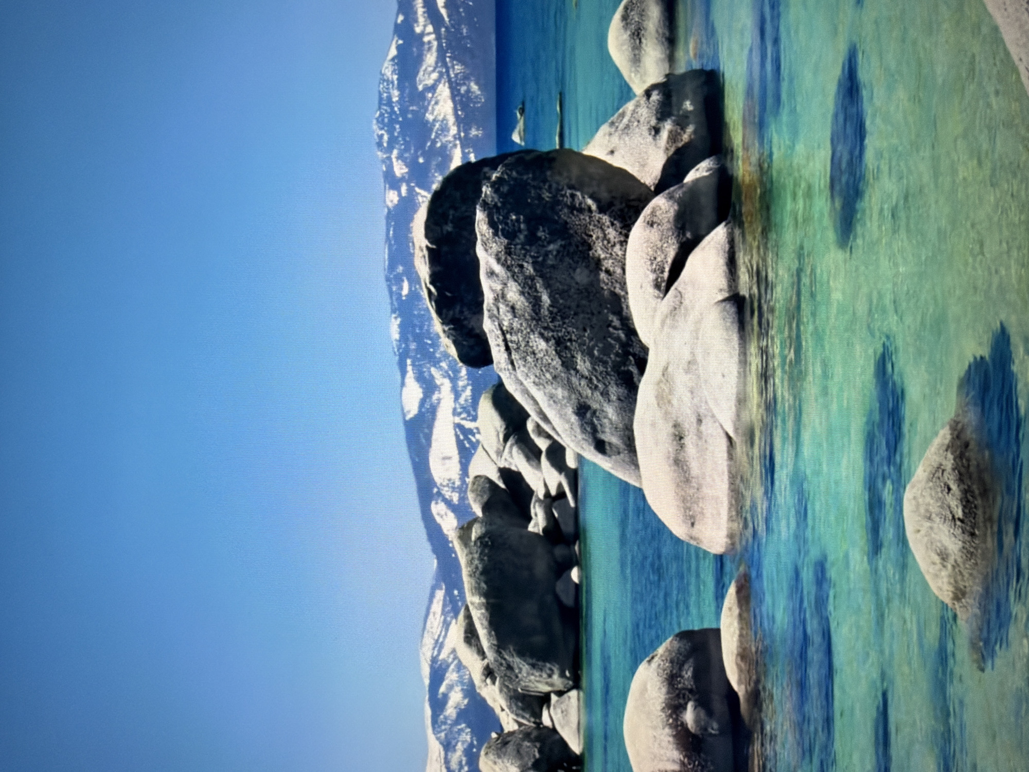 The image depicts a scenic view of a clear, turquoise lake with numerous large, smooth, grey boulders scattered in and around the water. In the background, snow-capped mountains rise against a vibrant blue sky. The water is shallow enough in some areas to see the lakebed and shadows cast by the boulders. There are no people visible in the image. The environment suggests a natural, outdoor setting, likely a lake in a mountainous region. This scene is not located in the city of Kyiv, Ukraine.