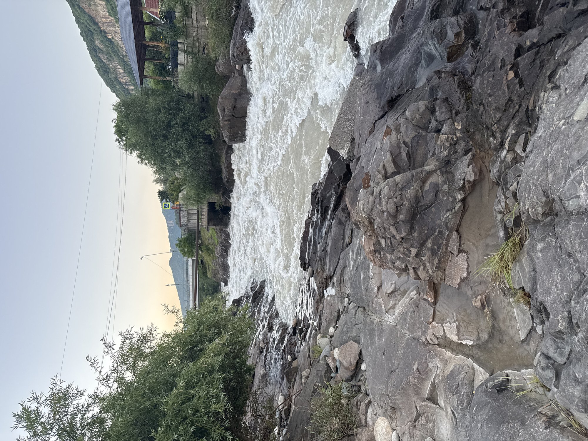 The image depicts a scenic view of a rushing river flowing through a rocky gorge in Kamennomostsky, Russia. The river, with its white, turbulent water, dominates the upper portion of the frame. The banks are lined with large, dark gray rocks, some appearing wet from the spray of the water, and interspersed with patches of green vegetation.
To the left of the river, a steep, tree-covered mountainside rises towards the sky. In the background, there are signs of human development, including buildings with dark roofs and a bridge crossing the river. Further in the distance, a street light and power lines are visible against the pale, hazy sky, suggesting either early morning or late evening. The sky itself is a soft, gradient hue, transitioning from a light blue at the top to a warm, peachy color near the horizon, indicative of sunrise or sunset.
The overall mood is one of natural beauty and raw power, with the dynamic flow of the river contrasting with the solid, ancient appearance of the rocks and mountains. There is no visible text in the image.