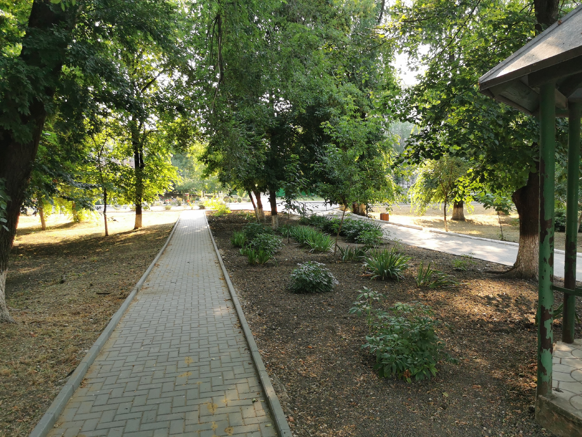 The image depicts a paved walkway lined with trees and green shrubbery in an outdoor setting, likely a park or garden. The walkway is made of grey interlocking pavers and runs from the foreground into the distance, curving slightly to the right. To the left of the walkway is a grassy area with some dry leaves and a few trees. To the right of the walkway, there is a flowerbed with various green plants, including some spiky, grass-like foliage and a rounded bush. Further to the right, partially visible in the foreground, is a weathered, green-painted structure, possibly a gazebo or shelter, with visible supporting posts and a roof. Sunlight filters through the trees, casting dappled shadows on the ground and walkway. The overall impression is a well-maintained outdoor space on a sunny day.