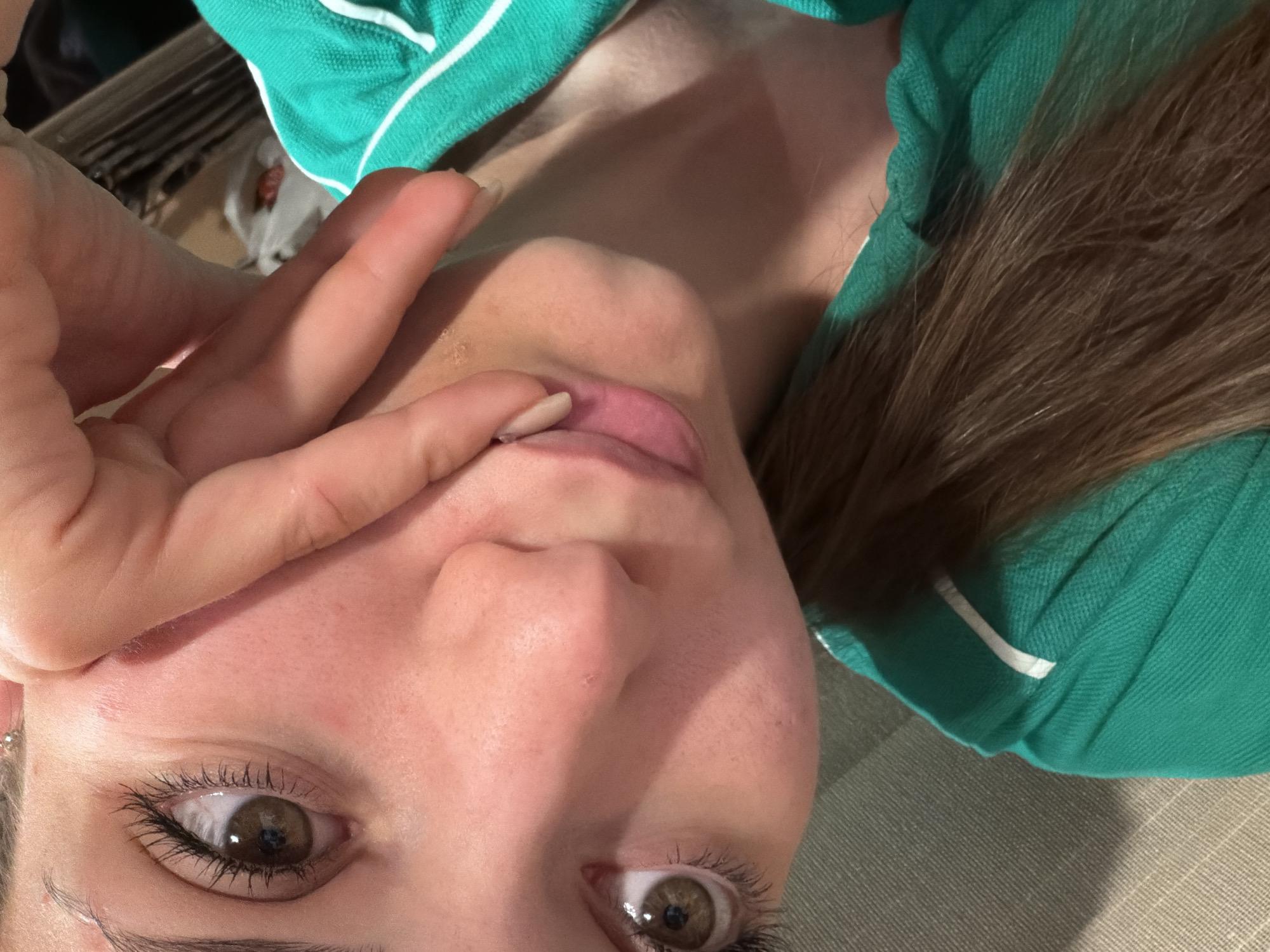 A close-up, low-angle selfie-style shot features a fair-skinned person with brown hair wearing a green and white collared shirt. The person's left hand is raised, partially obscuring their face with fingers near the eyes. The focus is on the face, particularly the eyes and nose, showing detailed skin texture. The background is blurred and neutral in tone. The location in Buhovo, Bulgaria, is not discernible.