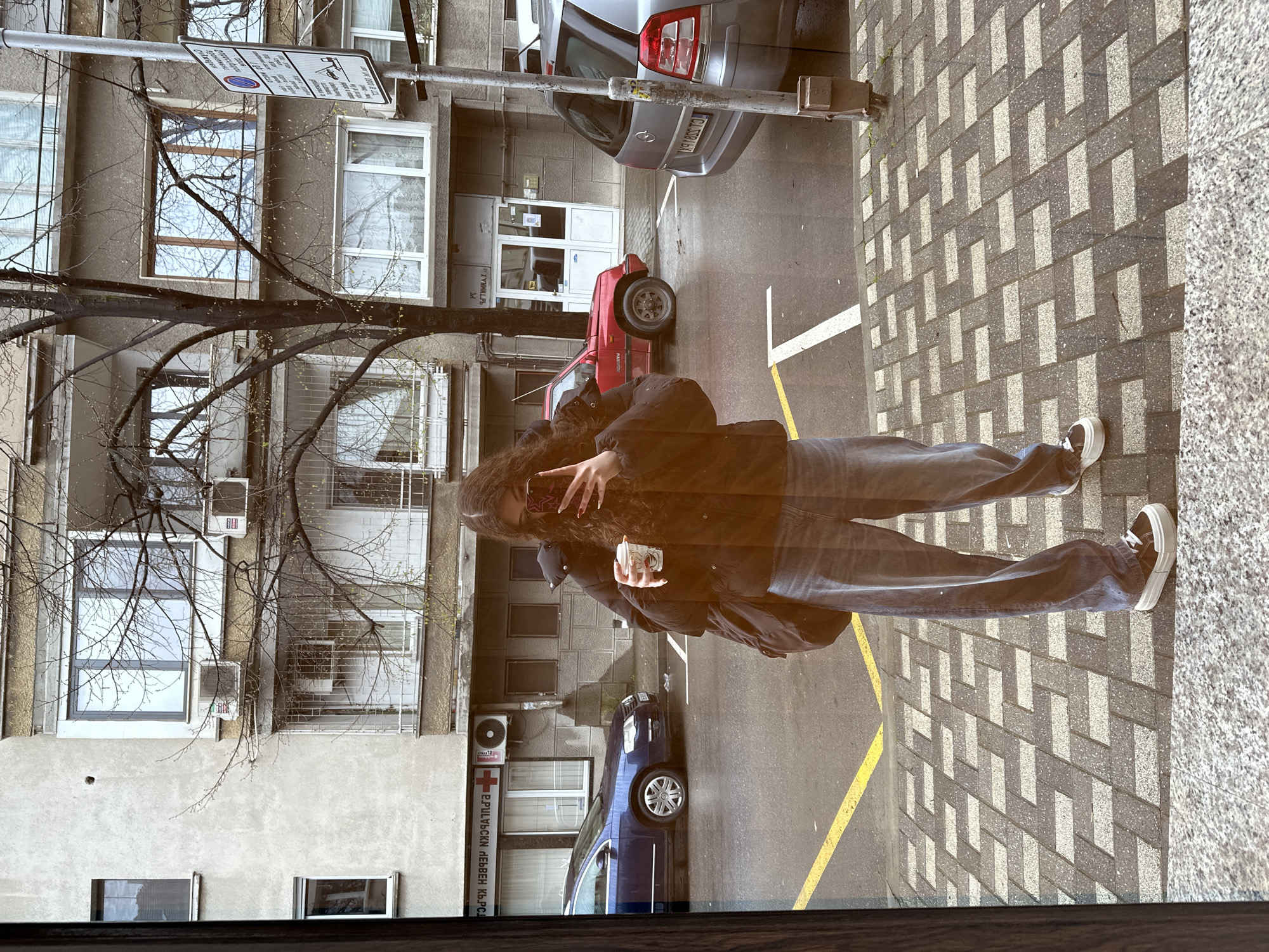 A woman is taking a mirrored selfie outside of a building with a sign written in Bulgarian, which translates to "First Aid Medical Center." A tree with bare branches is in the foreground. The street is wet, and several cars are parked. The sidewalk is paved with gray rectangular stones. The location is likely in Stara Zagora, Bulgaria.