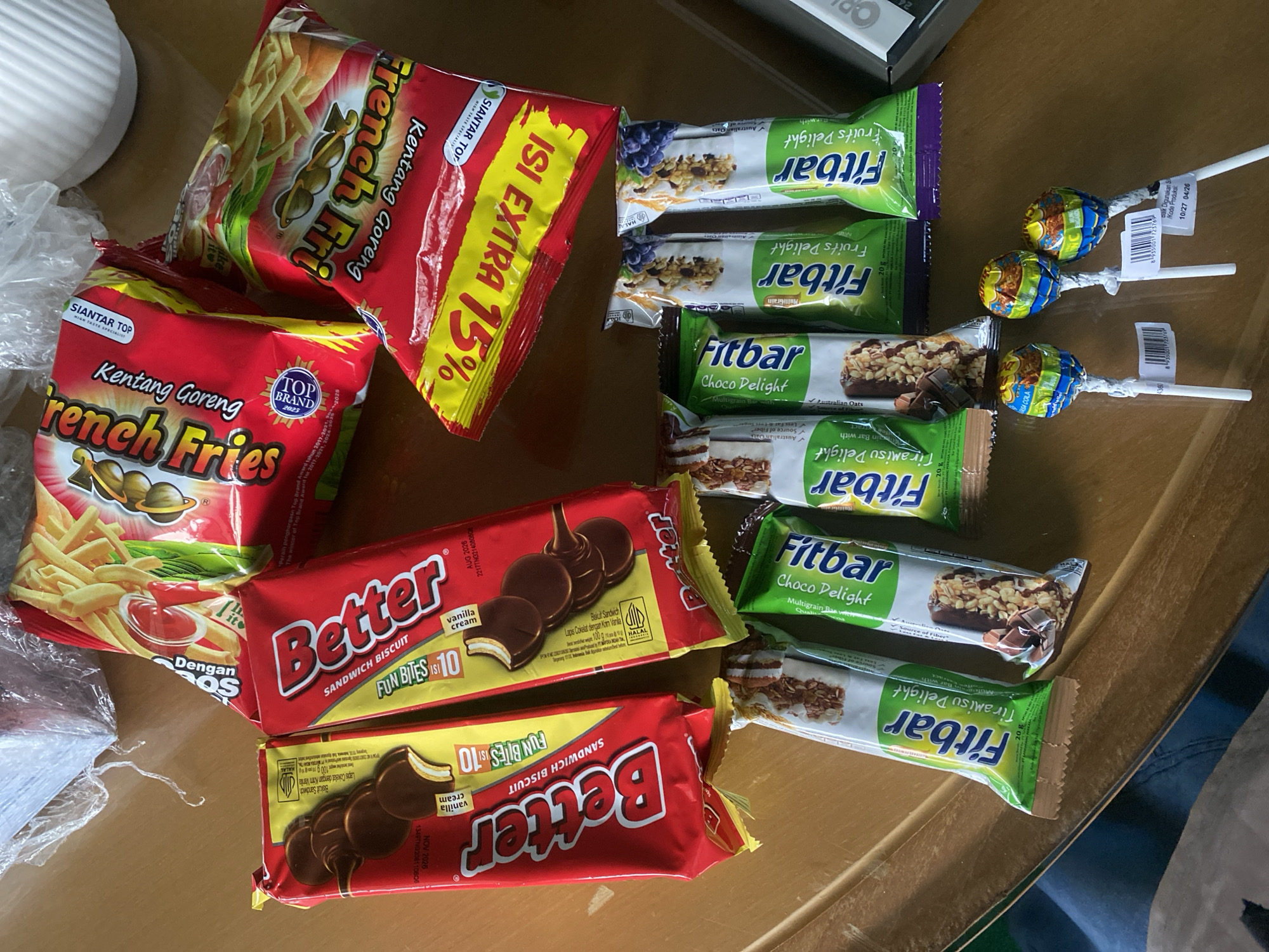 An overhead, slightly angled shot shows a collection of snack foods arranged on a wooden surface, likely a table. The snacks include two red and yellow packaged "Better" brand sandwich biscuits with chocolate-covered cream filling, and two red packaged "French Fries" brand potato snacks. Below these, several "Fitbar" brand fruit and chocolate flavored snack bars are visible, some in green packaging and others in white with brown accents. Three lollipops with colorful wrappers are at the bottom of the frame, on white sticks. The scene appears to be an indoor setting, possibly a home or store, given the arrangement of food items. There are no people visible in the image. The lighting suggests a bright, possibly natural, light source. There is no specific indication of the location within the city of Banjarbaru, Indonesia, other than the presence of Indonesian product packaging.