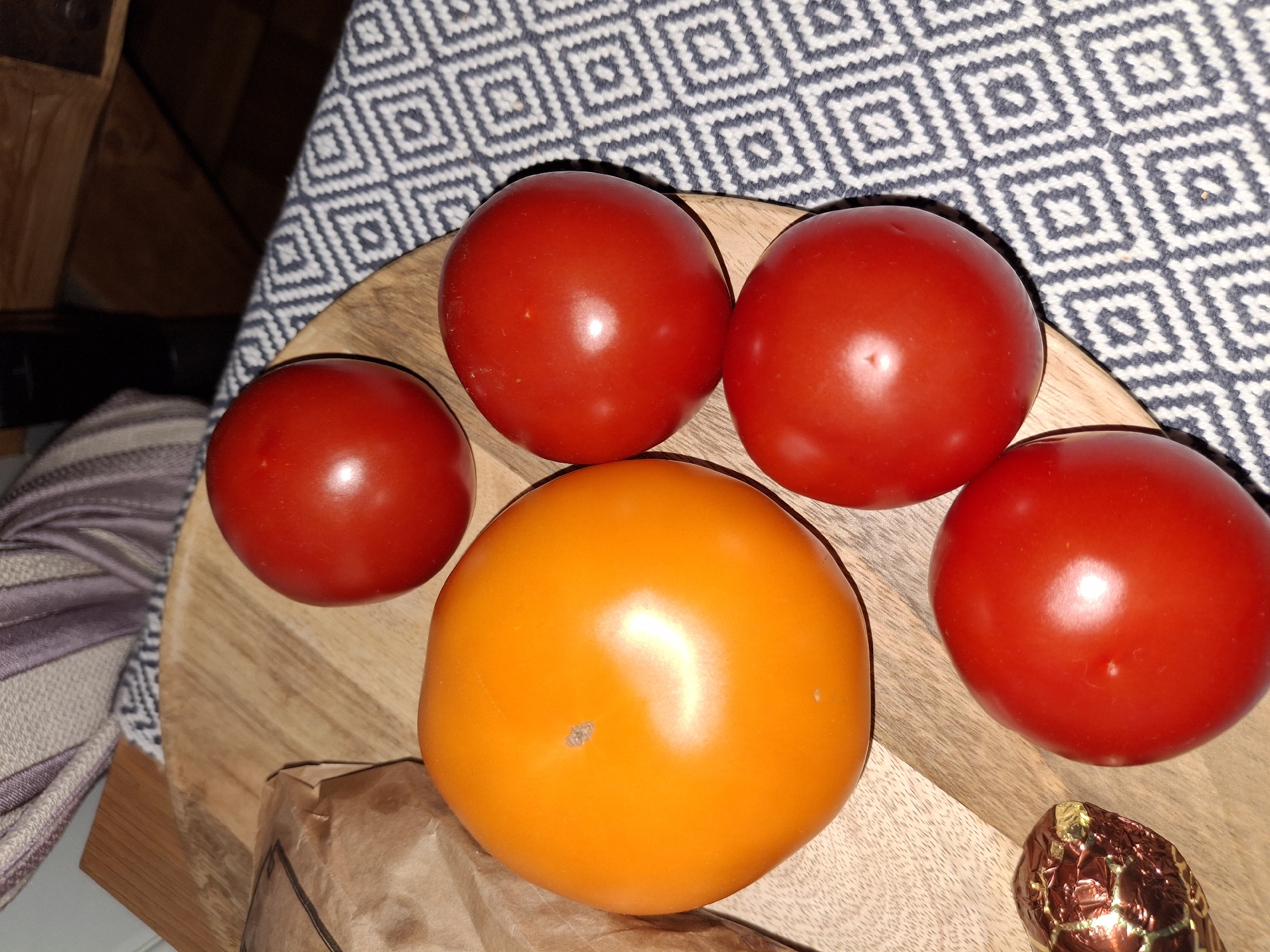 The image depicts a close-up, overhead view of several tomatoes arranged on a wooden board. The tomatoes vary in color, with one large, bright orange tomato dominating the left side of the frame, and four smaller, red tomatoes scattered around it. One of the red tomatoes is positioned above the orange one, another to its right, and two more below and to the right of the orange tomato. A small, crinkled, copper-colored object, possibly a piece of candy or a truffle, is visible to the lower left of the orange tomato. The wooden board has a visible grain pattern. The background consists of a textured, patterned cloth with a repeating diamond design in grey and white, and a portion of what appears to be a wooden surface, possibly a table or shelf, is visible at the top. The lighting suggests an indoor setting, with highlights reflecting off the smooth surfaces of the tomatoes. The specific location within Åsgårdstrand, Norway, is not discernible from the provided image.
