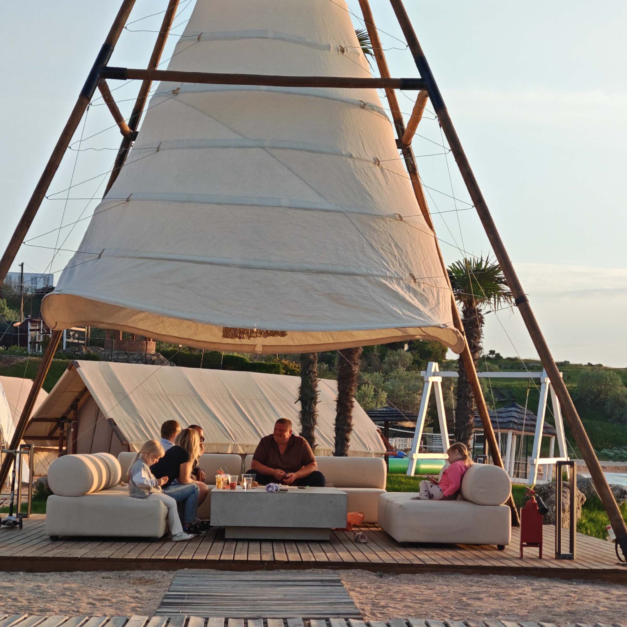 The image depicts a gathering of people on an outdoor deck, possibly in a resort or recreational area. The scene is characterized by a large, sail-like structure overhead, supported by a wooden framework. This structure provides shade or shelter.
There are five people visible in the image. A man in a brown shirt is seated on a white sofa along with two women and a young girl, who seem to be engaged in conversation. Another young girl is seated on another section of the same sofa, at the other end. A low, concrete table sits in front of the sofa, with drinks placed on top.
Behind the deck, there are tents. Palm trees stand in the background. A swing set is positioned near the edge of the deck. A red fire extinguisher is also visible near the edge of the deck.
The deck is made of wooden planks, leading to a beach-like area with sand. The location is possibly in the city of городское поселение Щелкино, Ukraine. The overall atmosphere suggests a relaxed and casual setting.