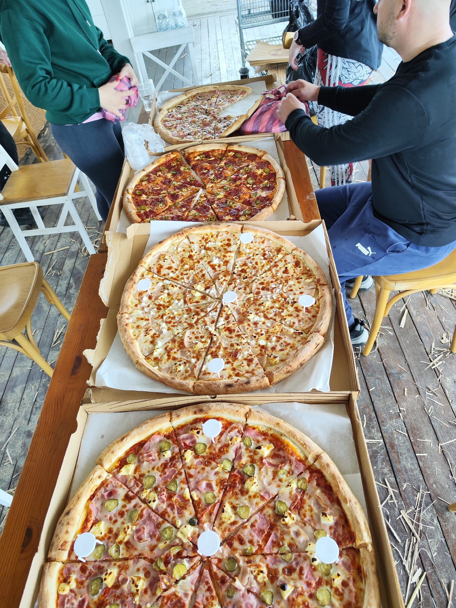 A high-angle shot captures a scene involving four large pizzas served from cardboard boxes on a wooden table. Two people are partially visible, with one seated on a yellow chair, wearing a long-sleeved black shirt and blue Puma sweatpants, and the other standing, clad in a green hooded sweatshirt and dark leggings. They appear to be interacting with the pizzas and some plastic bags.
The pizzas vary in topping composition. One features pepperoni and green peppers, another has what appears to be bacon and a light-colored cheese, a third has pepperoni, and the last is topped with ham and pickles. Each pizza is pre-sliced and has small, white plastic separators between the slices.
The setting seems to be outdoors, potentially a patio or deck, with straw or hay scattered on the wooden flooring. There are wooden chairs, a metal shopping cart, and white cabinets or a wall in the background. Based on the items visible, it could be a fast food eatery in Burgas, Bulgaria.