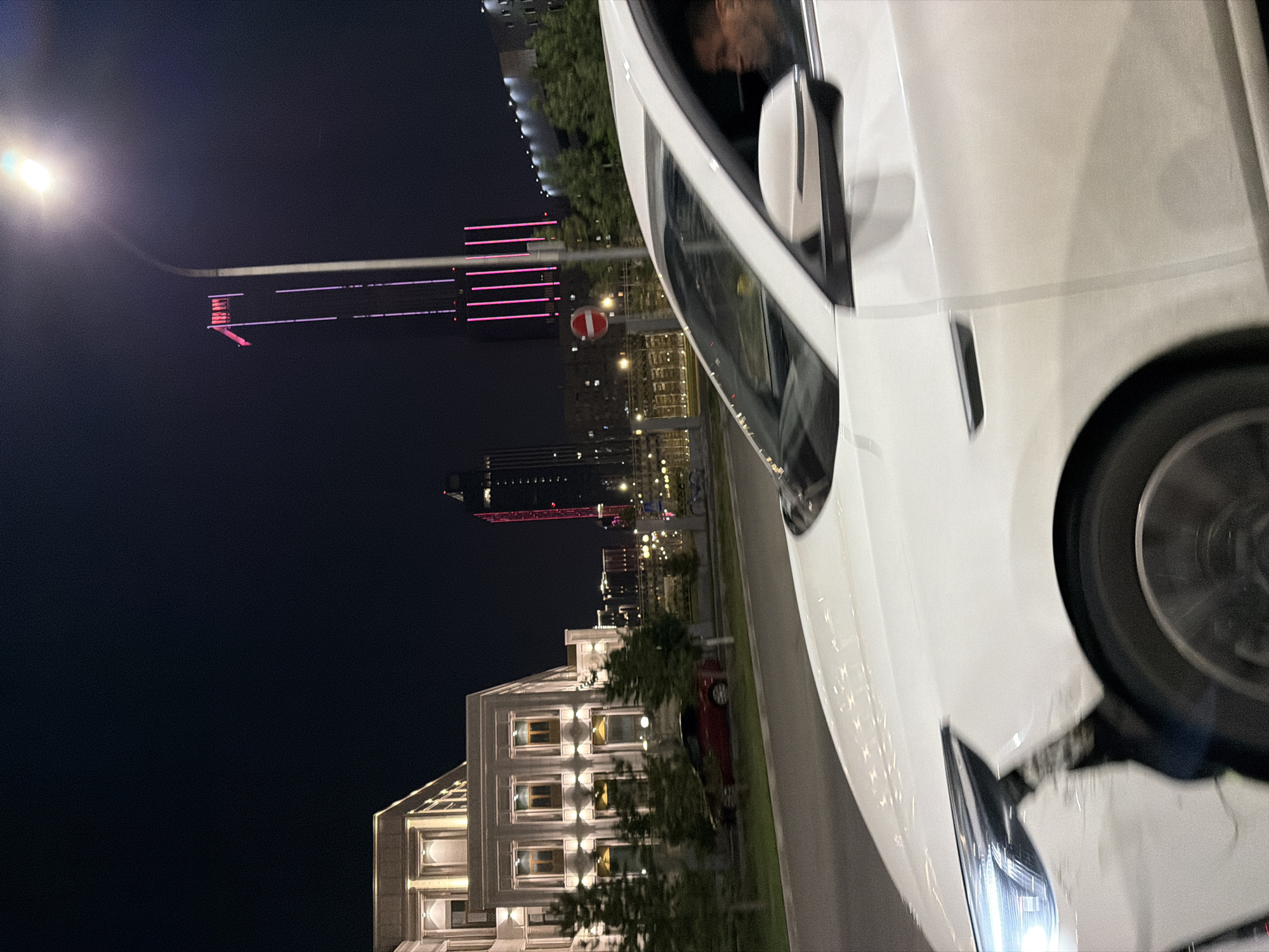 The image shows a nighttime scene in Astana, Kazakhstan, focusing on a white car in the foreground. The car, seen from the side, has damage to its front right fender and bumper. The driver, a man, is visible in the driver's seat, looking towards the right.
In the background, a modern building with illuminated windows is on the left. Further back, a tall skyscraper with pink and red lights is a prominent feature, with a streetlamp casting light over it. Other city lights and buildings are visible in the distance. A red car is parked on the side of the road near the modern building. The overall environment is an urban street at night.