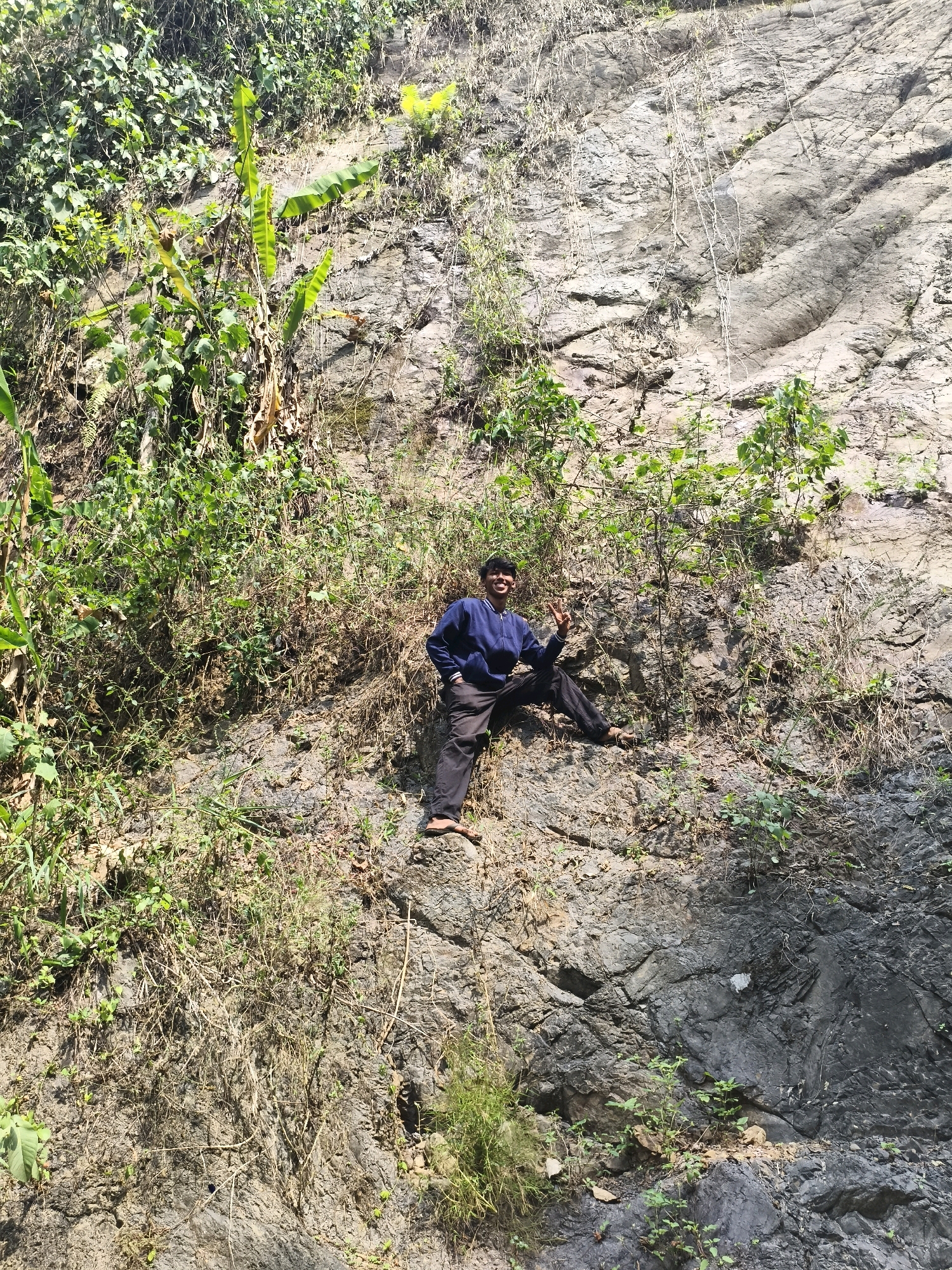 A full, eye-level shot shows a young man posing on a rocky cliff face in Babakan Madang, Indonesia. The man is positioned in the center-left of the frame, seated with his legs outstretched. He is wearing a dark blue hooded sweatshirt and dark pants. His right hand is raised, forming a peace sign, and he is smiling at the camera. The cliff face is covered in a mixture of rocks, dry vegetation, and green plants. Banana trees are visible on the left side of the image. The lighting suggests it is daytime, with shadows cast on the rocks, indicating the sun is high. The overall scene depicts an outdoor adventure in a natural, rugged environment.