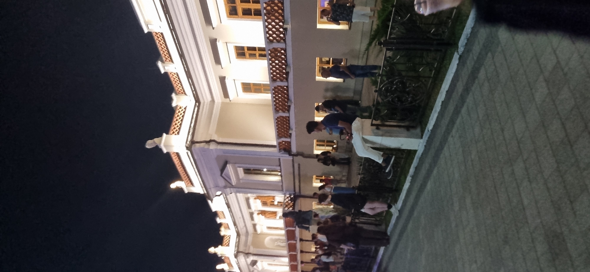 The image depicts an evening scene outside a large, ornate building with classical architectural elements. The building is illuminated by spotlights, highlighting its white facade, decorative cornices, and a patterned balcony railing. Several windows are visible on the upper floors, some of which are lit from within.
In the foreground, a paved walkway runs along a decorative metal fence. A group of people are gathered on either side of the walkway and near the fence. One individual is sitting on a white concrete post, wearing white pants and a dark shirt, and appears to be holding a remote control. Other individuals are standing and talking, some leaning against the fence.
In the mid-ground, a dark statue on a pedestal is visible in front of the building, partially obscured by the fence and people. The background is a dark night sky.
The scene is located in Feodosia, Ukraine. The building's architectural style and the presence of a statue suggest it could be a historical or cultural institution, such as a museum or administrative building. The gathering of people indicates an event or a popular public space. The lighting and attire of the individuals suggest a mild evening.