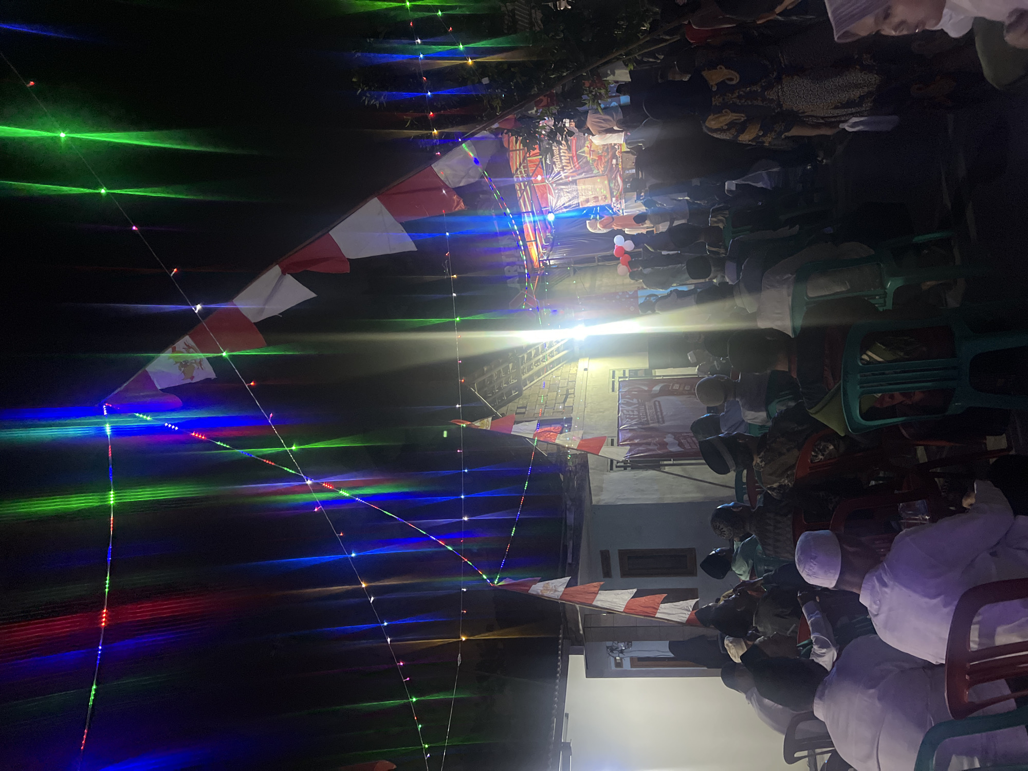 The image depicts an outdoor gathering at night in Klapanunggal, Indonesia, likely a festival or event. The scene is illuminated by strings of colorful lights, primarily red, green, and blue, which cast vibrant beams. Several Indonesian flags, red and white, are hung prominently, suggesting a patriotic or celebratory occasion.
In the background, a stage is set up with lighting and a banner that appears to read "FESTIVAL BERSATU." People are gathered, with some seated on chairs in an audience area and others standing near the stage. The audience members are predominantly men wearing traditional Islamic attire, such as peci (caps) and white shirts. A few women are also visible, some wearing headscarves.
The environment includes modest buildings and trees, indicating a semi-urban or rural setting. The overall atmosphere appears festive and communal.