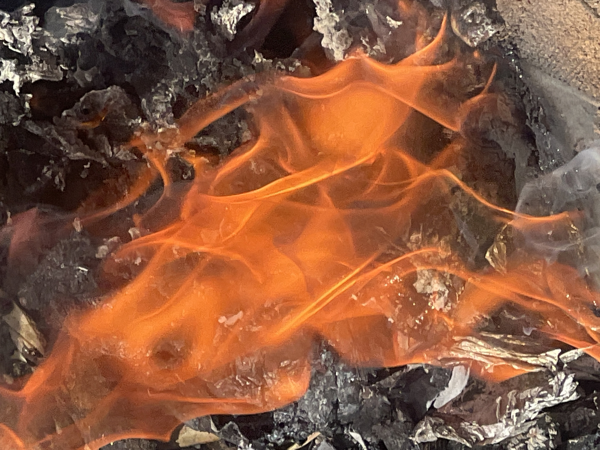 The image depicts a close-up of burning material. Flames, predominantly orange, rise from a bed of dark gray and black ash and partially combusted debris. Some white flecks are visible, possibly unburned particles. The specific location in Gura Top, Nigeria, is not discernible from the image. No people or interactions are visible.