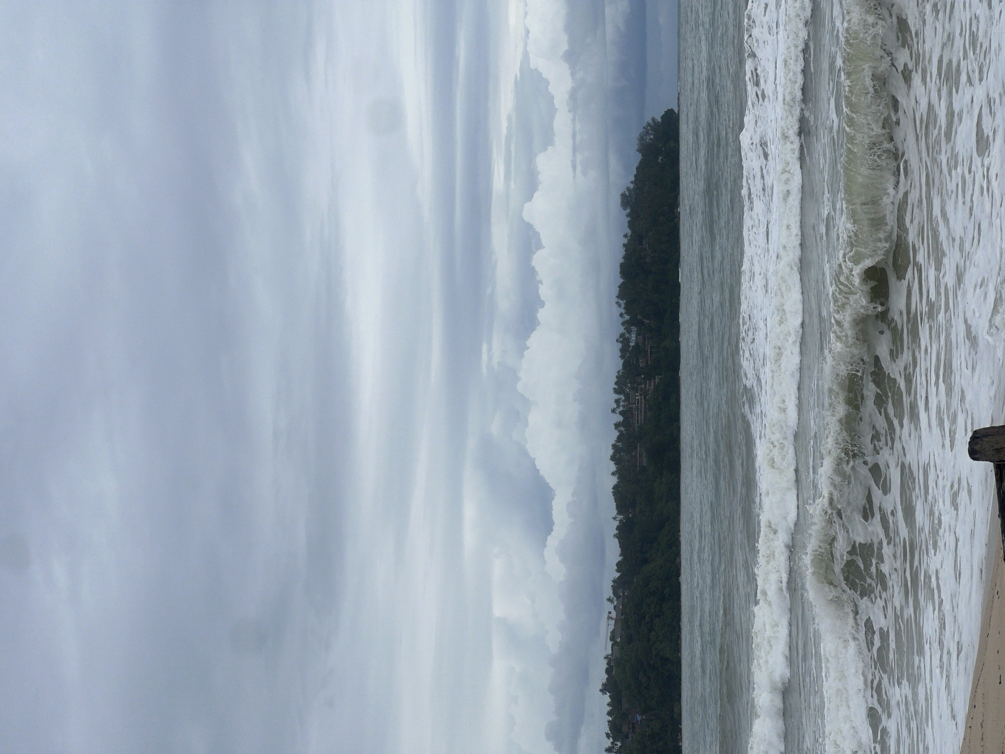 This is a photograph of a beach scene on a cloudy day. In the foreground, there are waves breaking on the shore. The water is a muted gray-green color, and the waves are frothy and white. A small section of a wooden post is visible in the lower right corner of the image. In the middle ground, there is a body of water, likely the ocean, and beyond that, a lush green hillside covered in trees and vegetation. Several buildings are visible nestled into the hillside, suggesting a resort or residential area. The background is dominated by a gray, overcast sky with layered clouds. The overall impression is one of a tranquil, natural setting under a somber sky.