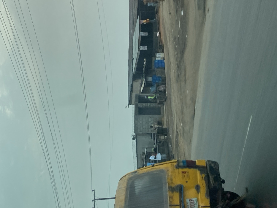 A roadside view shows a weathered yellow auto-rickshaw. Next to it is a row of modest structures, likely shops or workshops, with corrugated metal roofs and cinder block walls. Electrical wires crisscross the sky, adding to the utilitarian feel of the scene. The road is paved and appears slightly wet, contributing to the subdued light under an overcast sky. People are seen near the shops, engaged in activities that suggest a bustling yet commonplace street.