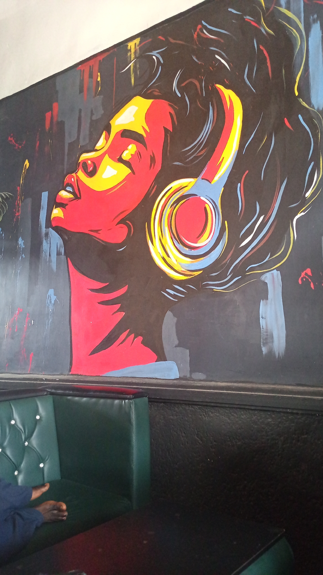 This image captures a vibrant mural painted on a wall, depicting a stylized portrait of a person wearing headphones. The artwork features bold colors, with a predominantly red and yellow face against a dark background. Streaks of red, blue, and white paint add a dynamic element to the mural, suggesting movement or sound waves emanating from the headphones. The headphones themselves are rendered in concentric circles of yellow, red, and blue.
In the foreground, a section of a dark green booth with tufted upholstery is visible. A person's bare feet and lower legs, clad in dark blue trousers, are resting on the booth's seating. The overall scene appears to be an indoor setting, possibly a restaurant, bar, or lounge, given the booth seating and the presence of a dark tabletop in the lower right corner. The lighting suggests it could be daytime, but the mood is amplified by the dramatic artwork. No text is visible in the image. The location is identified as Howayi, Nigeria.