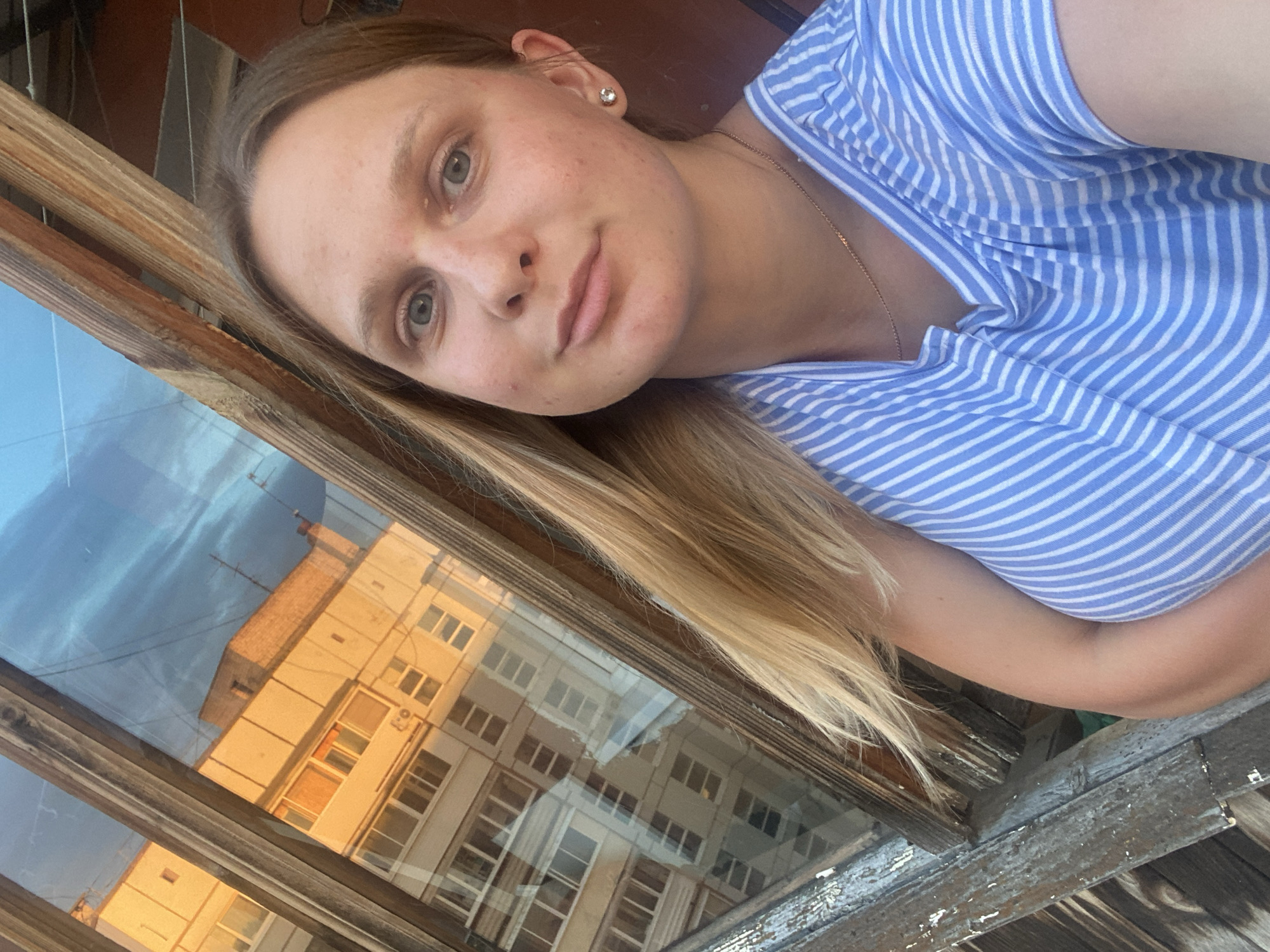 A young Caucasian woman with blonde hair and blue eyes is taking a selfie in front of a window. She is wearing a blue and white striped t-shirt and a gold necklace. Her right arm is bent, and she is looking at the camera with a slight smile. The window shows a reflection of buildings and a blue sky. The woman appears to be on a balcony or near a window in an urban setting, possibly in the city of Chaikovsky, Russia, given the architecture of the reflected buildings. The wooden window frame shows signs of weathering.