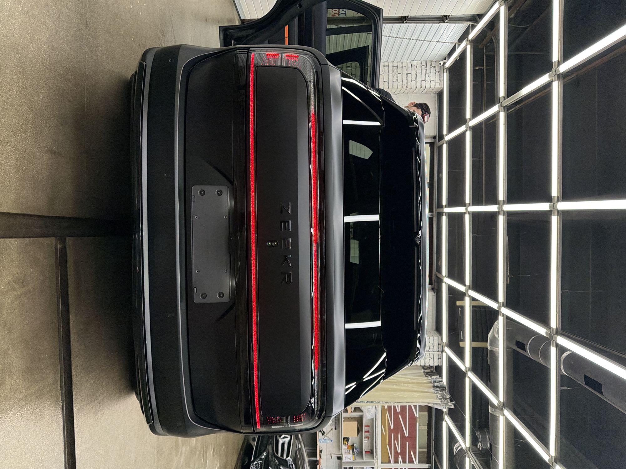 The image shows the rear of a black Zeekr car in a garage or workshop setting. The car is parked with its hatchback open. A person is visible inside the car near the open door. The garage is illuminated by a grid of fluorescent lights. The environment around the car includes elements suggesting an automotive repair or detailing shop, such as shelving and equipment in the background. There is no clear evidence to confirm the exact location is in Krasnoyarsk, Russia, but the assumption can be made based on available information.