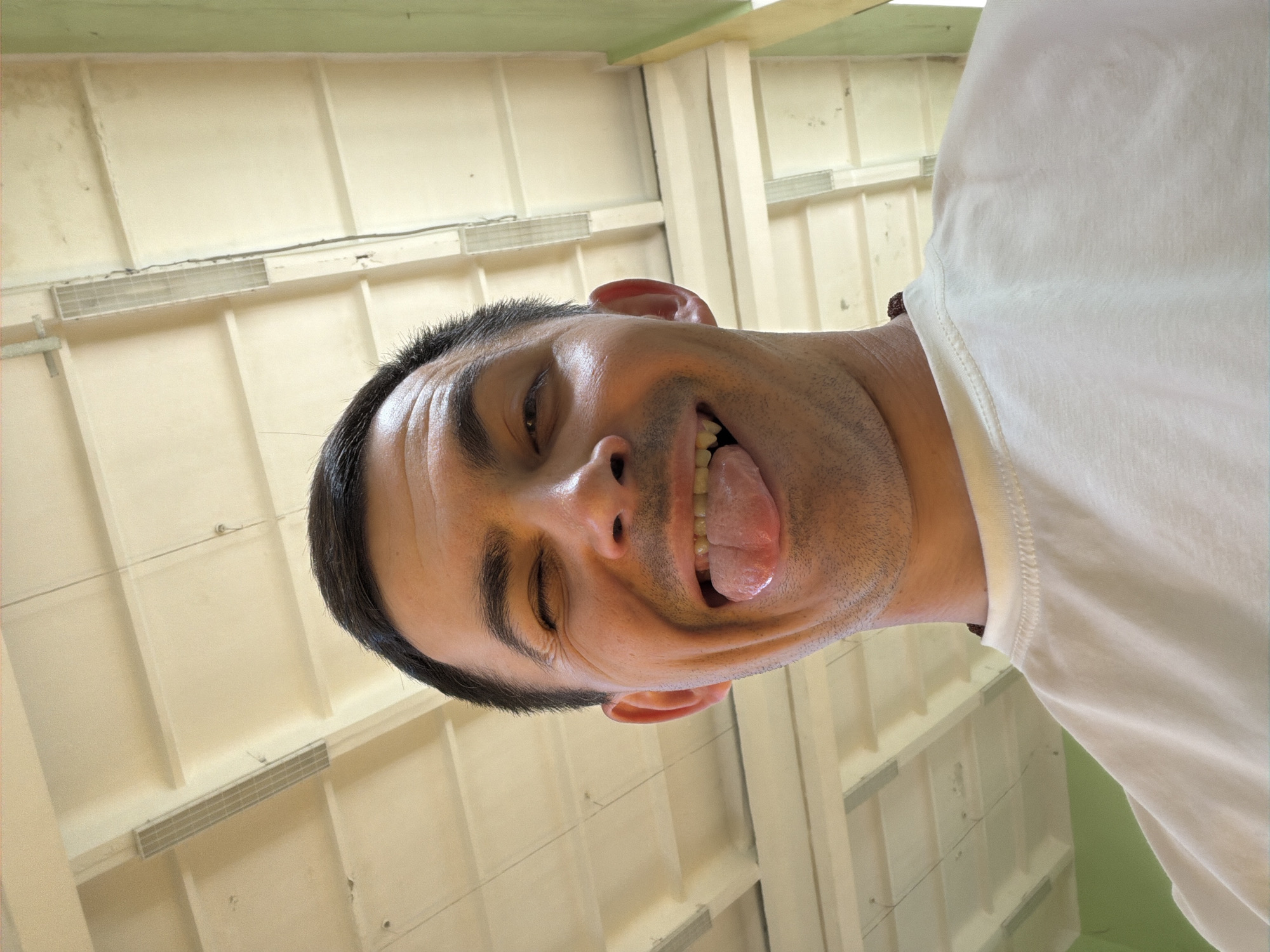 A man with dark hair and stubble, wearing a white shirt, is sticking his tongue out and winking. The background is a light beige or cream-colored wall with vertical and horizontal lines, possibly indicating a ceiling or interior structure. This image was not taken in Izhevsk, Russia.