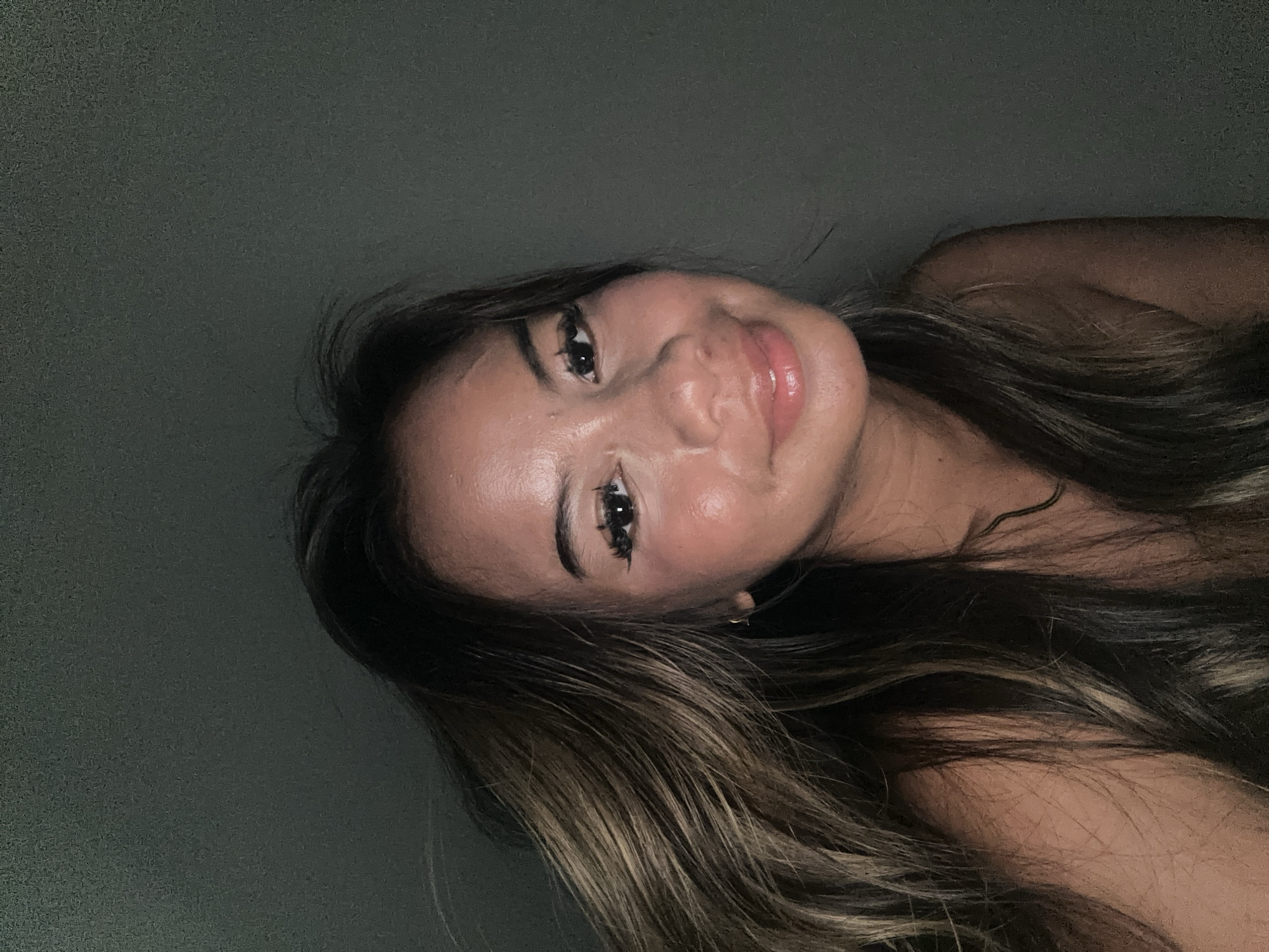 A medium close-up shot of a woman with long, wavy, brown hair. She has fair skin, dark eyes, and full lips. She appears to be wearing makeup, including eyeliner and lip gloss. The background is a solid, dark gray color. The image seems to be taken indoors. There is no information available in the image to determine the location is Sukawati, Indonesia.