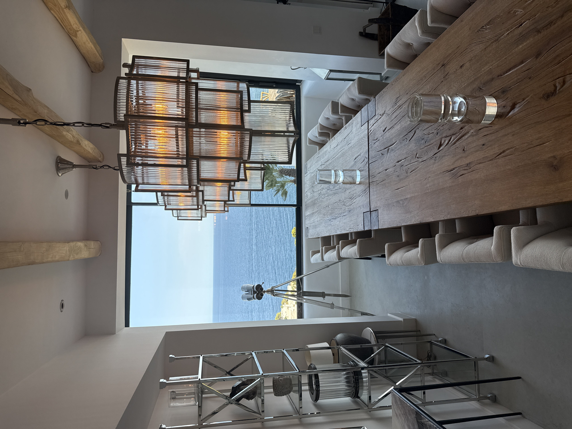 A spacious, well-lit dining room overlooks the Mediterranean Sea. The room features a large, rustic wooden dining table with upholstered chairs. A multi-tiered, Art Deco-inspired chandelier hangs above the table, casting a warm glow. To the left, a silver, multi-shelf étagère displays decorative objects. A pair of binoculars on a tripod stands near the window, pointing towards the sea. The window offers a panoramic view of the ocean and a rocky coastline. The room appears to be located in Santa Eulària des Riu, Spain, given the architectural style, natural light, and coastal scenery. The overall atmosphere is one of sophisticated, relaxed dining with a stunning natural backdrop.