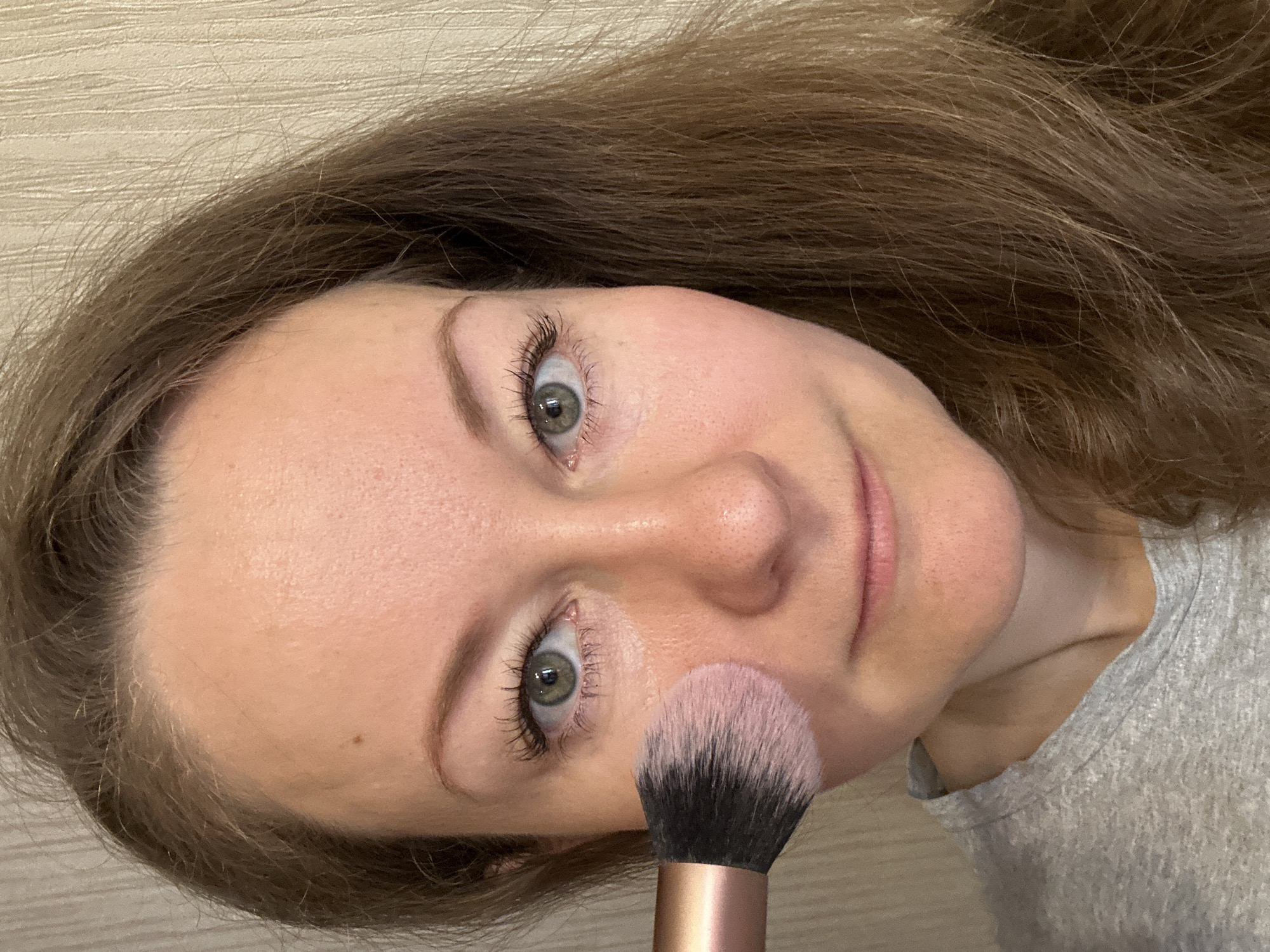 A woman is holding a makeup brush to her face. The brush has a black base with pink bristles. She has brown hair and light skin. She is wearing a gray shirt. The background is a beige wall. There is no indication that the photo was taken in Moscow, Russia.