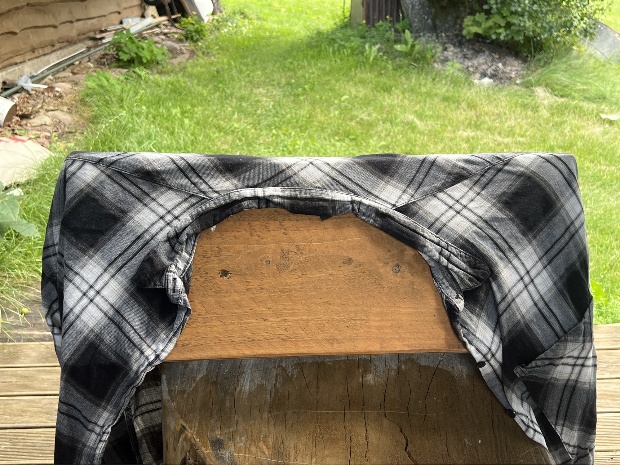 A close-up, overhead shot captures a black and gray plaid shirt draped over a wooden structure. The shirt is open, revealing a section of warm-toned wood. The background shows a blurred expanse of green grass and some out-of-focus foliage. The scene appears to be outdoors. There is no indication of the location being Norviliškės, Lithuania.