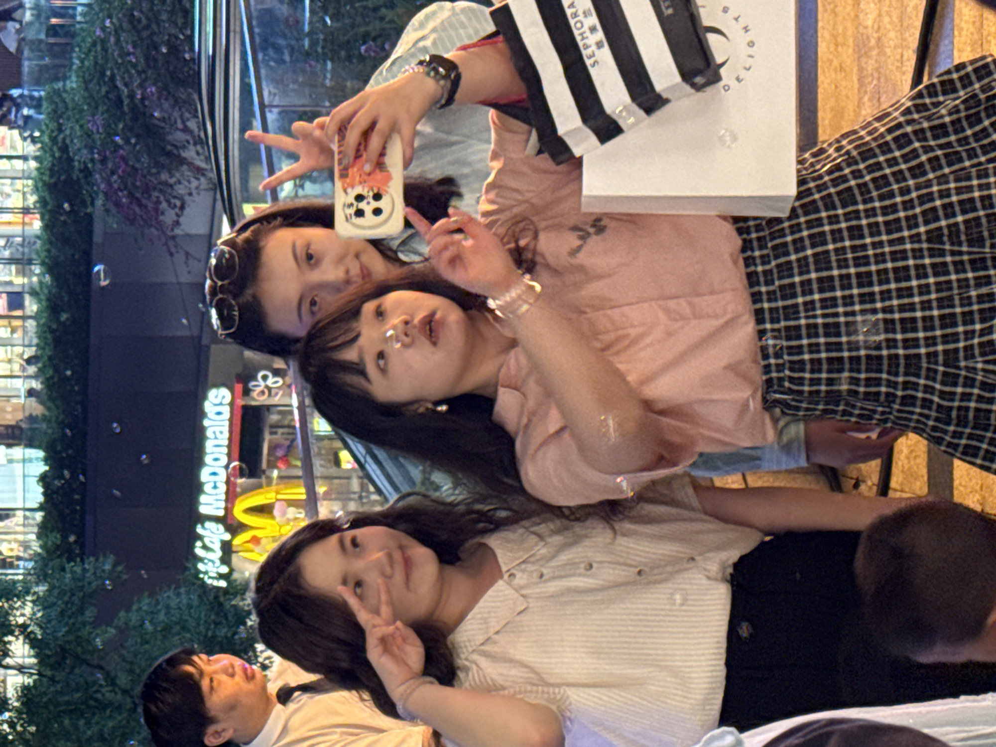 The image captures a group of people posing for a photograph. Three young women are visible in the foreground, making peace signs with their fingers, possibly for the camera. One woman is holding a phone.
The background shows a McDonald's restaurant. This indicates an urban environment, specifically in the Yuzhong District of China, given the mention of the location. The scene appears to be at night, based on the lighting and the presence of illuminated signs.
In the background, there are shops. One of the shopping bags is from Sephora. There is a variety of clothing styles among the women, including striped shirts and plaid patterns.