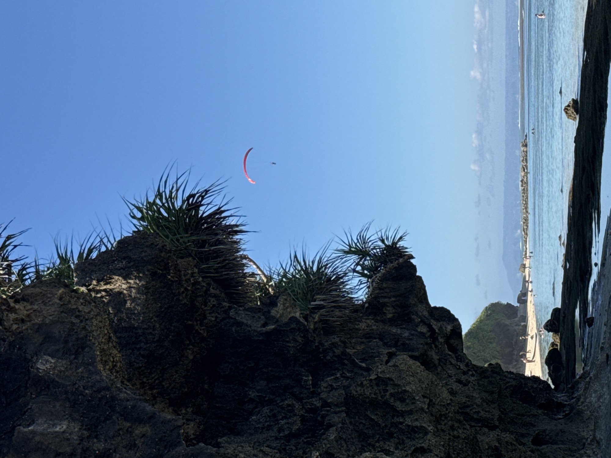 A red paraglider is visible in the clear blue sky above a tropical beach in Kuta Selatan, Indonesia. The paraglider, with a person attached, is soaring above the water. In the foreground, a large, dark rock formation juts out, covered in green vegetation. The beach itself is sandy, with several people visible walking and swimming in the turquoise water. In the distance, a breakwater or rocky structure extends into the sea, and mountains can be seen on the horizon under a hazy sky. The scene depicts a sunny day with calm waters and recreational activities.