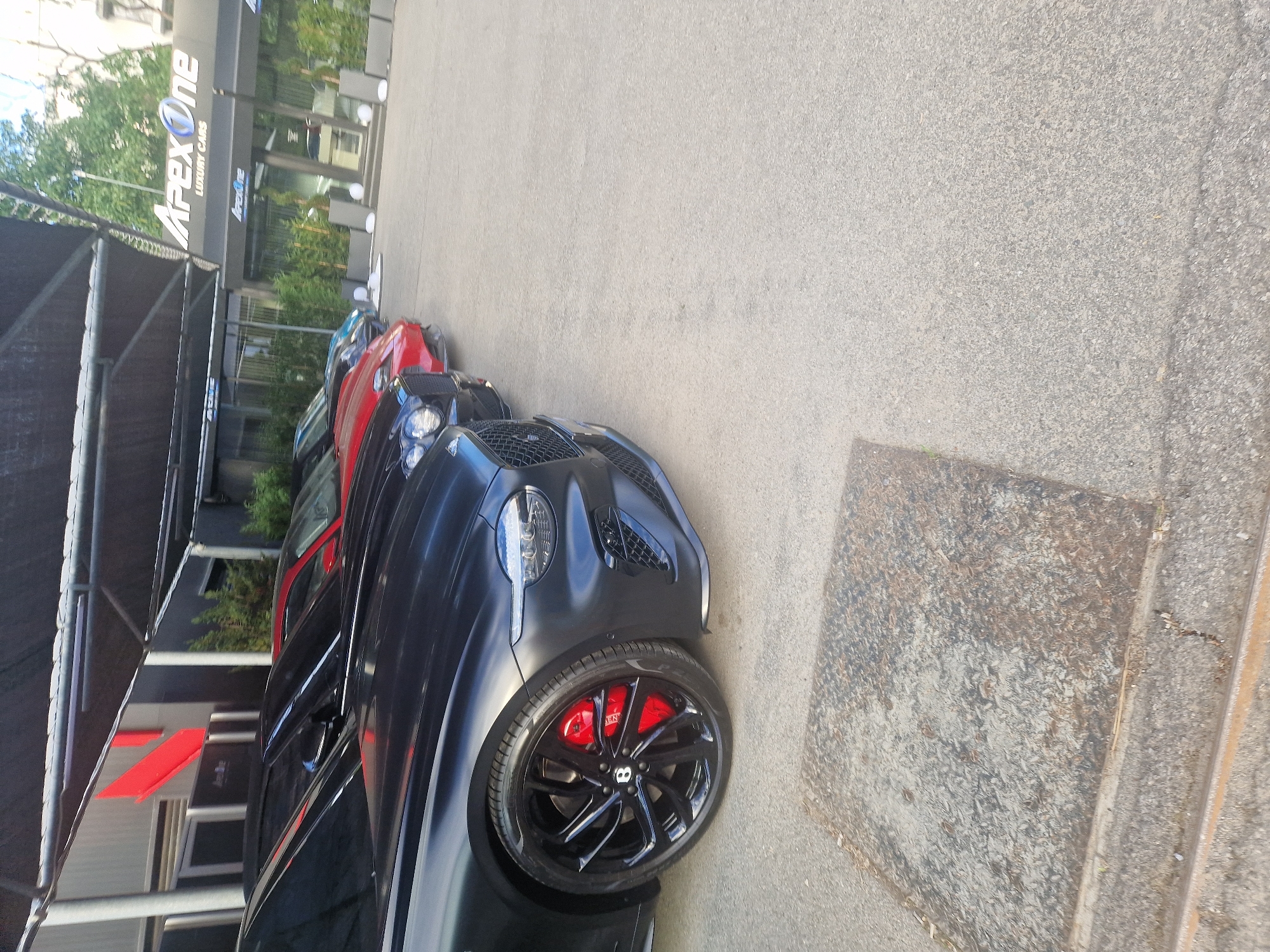 The image depicts several luxury cars parked in front of a building labeled "Apex One Luxury Cars." The cars are primarily dark-colored, with at least one red vehicle visible. The background includes greenery and a gray concrete ground. The location is in the city of Unknown, Unknown.