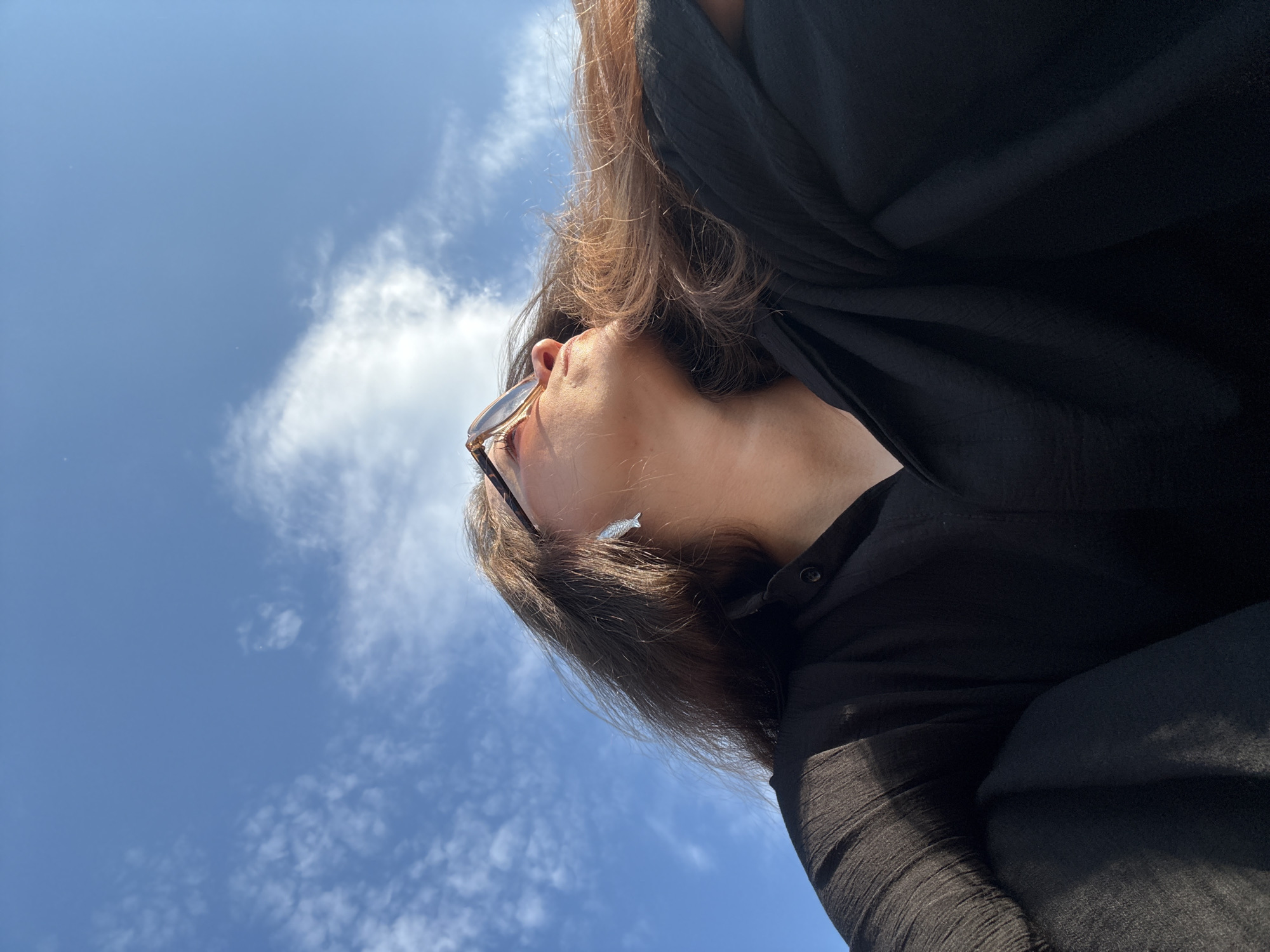 The image shows a low-angle shot of a person looking upwards towards a bright blue sky with scattered white clouds. The person has long, dark hair and is wearing sunglasses and a black collared shirt. A small, silver, fish-shaped earring is visible on their ear. The person's right arm is bent, and their legs are crossed in front of them. The background is dominated by the sky, with a hint of sunlight illuminating the person's face and hair. There is no discernible urban environment visible, and therefore, no specific location in the city of Chaikovsky, Russia can be determined from the image alone.