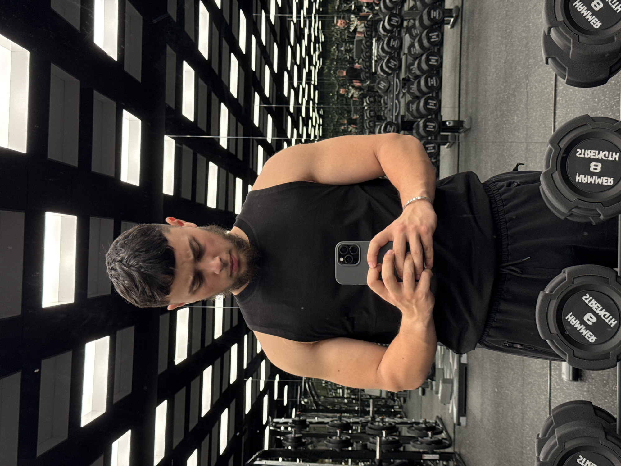 A young adult male, with dark hair styled in a modern cut and a short beard, is taking a selfie in a gym. He is wearing a black sleeveless shirt and black sweatpants. He is holding a grey smartphone in his hands, looking down at it. His left arm is bent, displaying his bicep, and he wears a silver bracelet on his left wrist. In the background, rows of weightlifting dumbbells are visible, neatly organized on racks. The gym ceiling features a grid of rectangular fluorescent lights. The scene is reflected in a large mirror, showing more gym equipment and a reflection of the man.
This image was captured inside a gym, likely in the city of Varna, Bulgaria, given the context provided. The specific gym cannot be identified further.