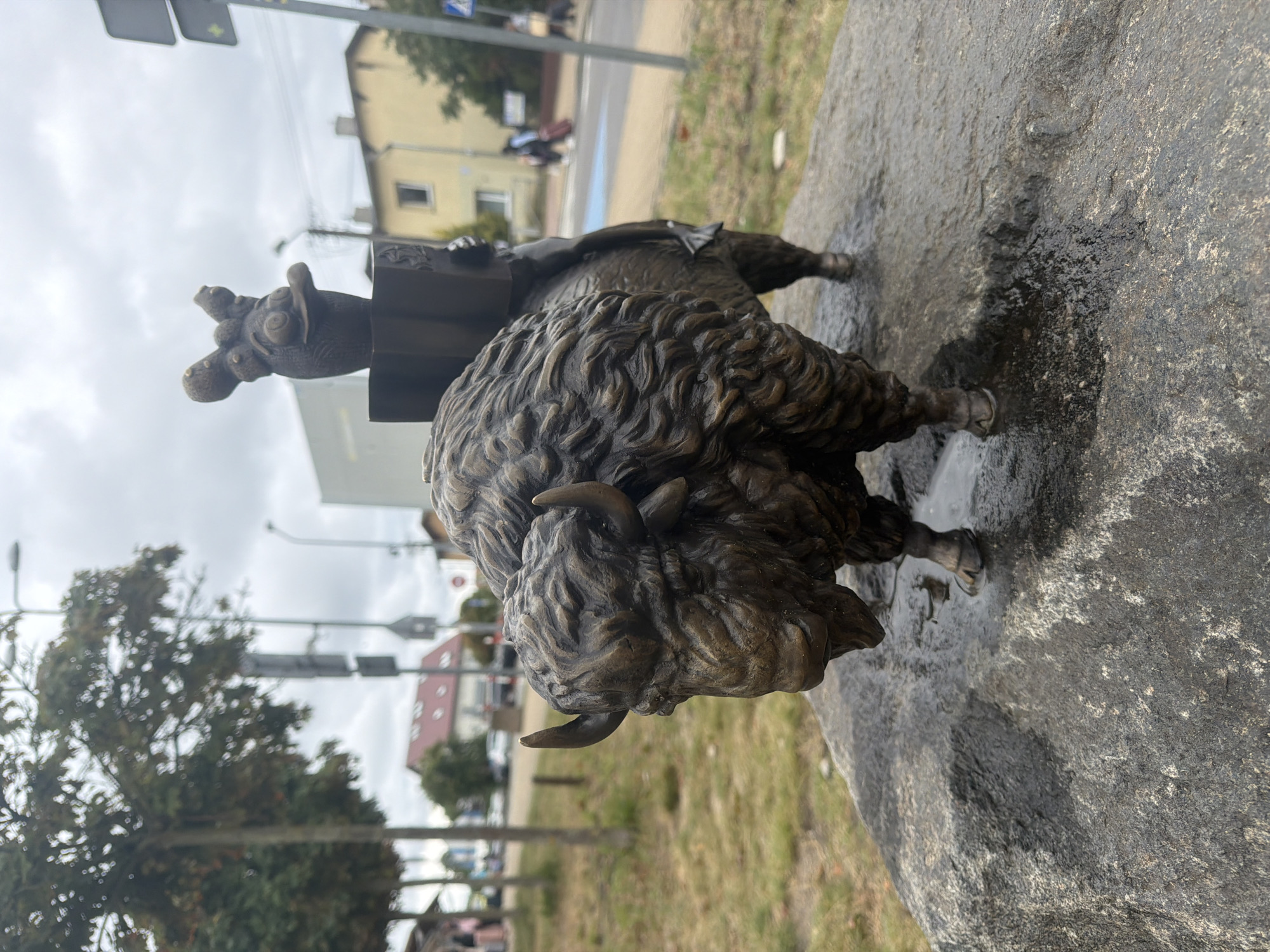 A bronze sculpture of a bison with a small, cartoonish figure riding on its back is positioned on a large, wet rock. The bison sculpture is detailed with textured fur and horns. The small figure atop the bison appears to be holding a box. The rock is wet, with puddles reflecting the overcast sky. In the background, there is a paved street with a pedestrian crossing and some blurry figures walking with luggage. Buildings, trees, and streetlights are also visible in the background, suggesting an urban or semi-urban environment. This sculpture is located in Międzyzdroje, Poland, specifically on the Promenade.