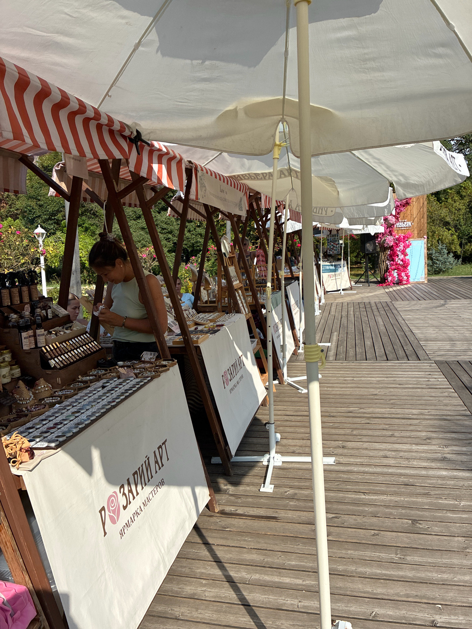 The image depicts an outdoor market scene, likely in Sochi, Russia, given the presence of Cyrillic text. The market features several stalls with white canopies and red and white striped awnings, set up on a wooden boardwalk. One stall prominently displays "РОЗАРІЙ АРТ" and "ЯРМАРКА МАСТЕРОВ" on a white banner, suggesting it's a craft fair. A woman with her hair tied back in a bun, wearing a light blue tank top, is seen behind one of the stalls, interacting with merchandise, possibly jewelry or small crafts. Another woman is partially visible behind her. The stalls are filled with various items, including what appear to be small bottles, jars, and rows of small containers, possibly holding beads or other craft supplies. In the background, trees and greenery are visible, along with a structure adorned with bright pink floral decorations. The wooden boardwalk extends into the background, indicating a public area, possibly a promenade or a park. The lighting suggests it is daytime, with sunlight casting shadows from the awnings and market structures.