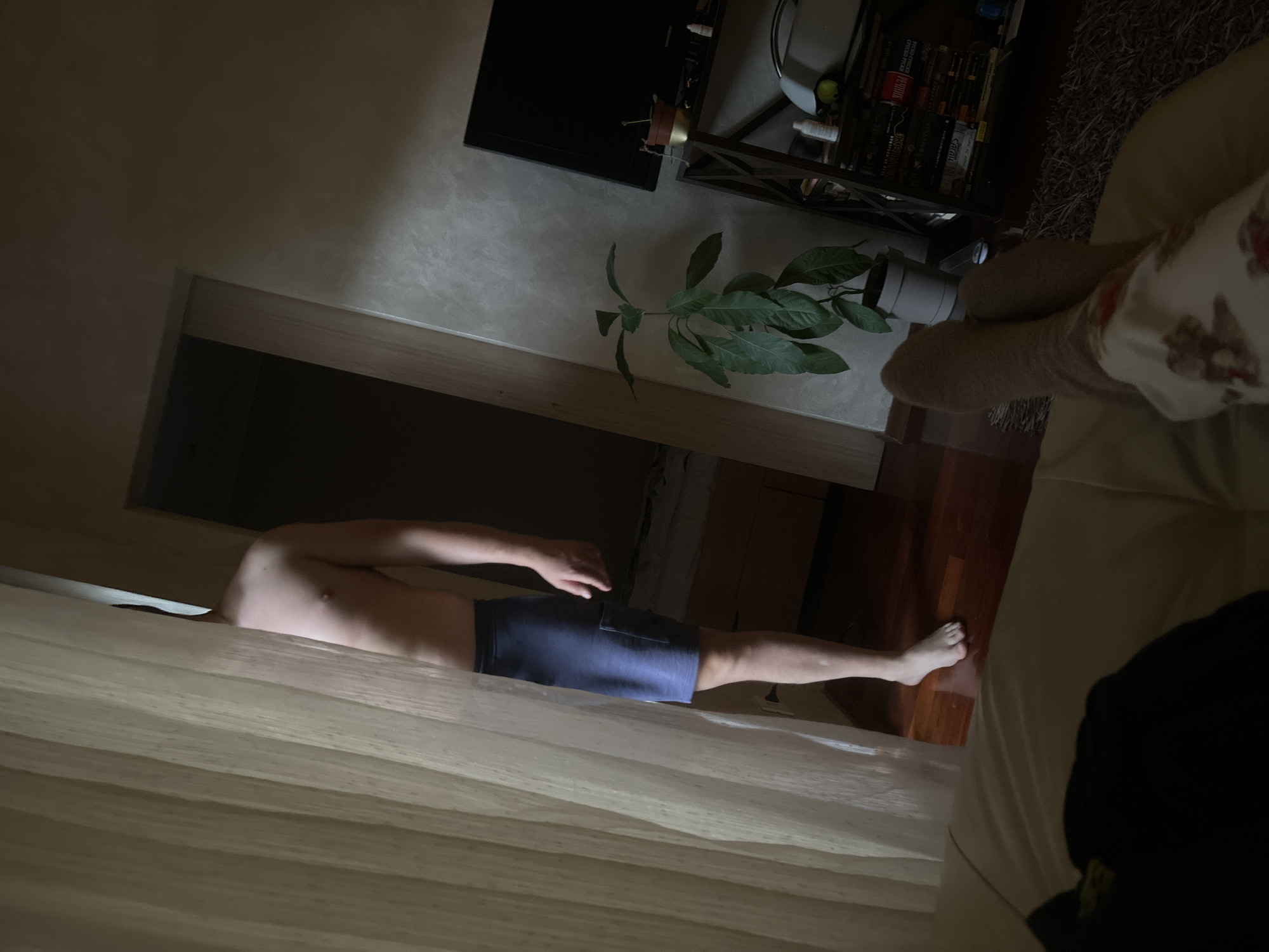A man is lying shirtless with blue shorts on a bed under a white curtain. His foot is touching a wood floor. In the background is a doorway, a plant in a grey pot on a table, a television, a black shelving unit with books, and a grey carpet. Part of a person's legs covered in patterned socks and blanket can be seen on the right. The location appears to be inside a residence, possibly in Budva, Montenegro.