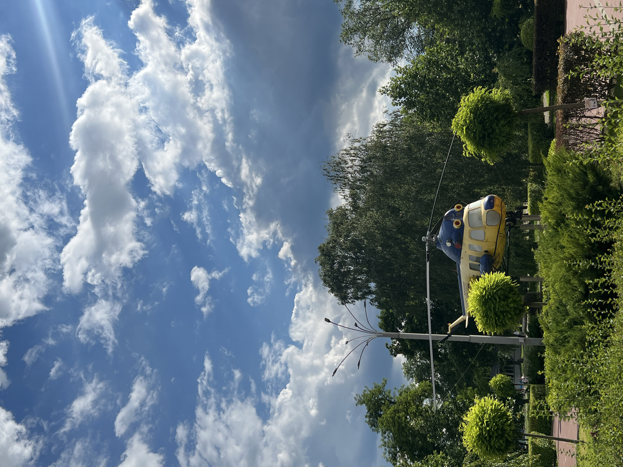 A yellow and blue helicopter is parked on a platform in a park. The helicopter is in the center of the image, and it is surrounded by lush green trees and bushes. The sky above is blue with white clouds. There is a lamppost to the left of the helicopter. The ground in front of the helicopter is a red paved path. The park appears to be well-maintained and is likely a public space. There are no people visible in the immediate vicinity of the helicopter.