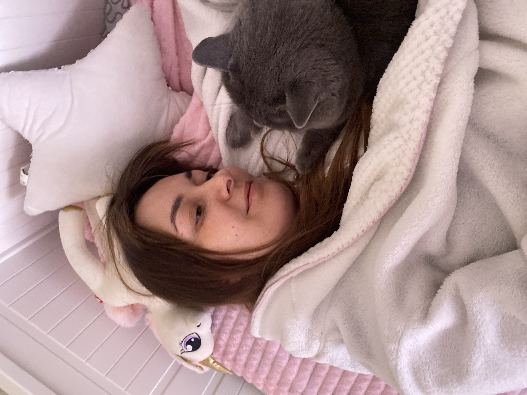 A young woman lies in a bed, partially covered by a fluffy white blanket with pink trim. Her head rests on a white star-shaped pillow, next to a white plush unicorn toy. A grey cat is positioned on her chest. The bed is likely in a room within a residence, but no specific details indicate a location in Votkinsk, Russia. The lighting is soft and diffused. There are no actions occurring, the woman is resting or sleeping with the cat on her.