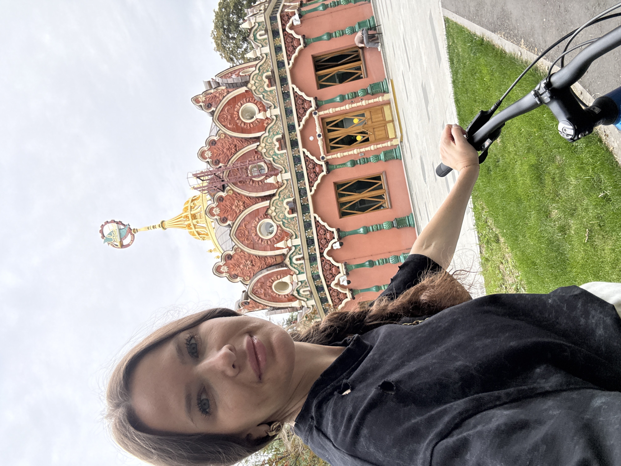 This is a selfie taken by a woman holding the handlebars of a bicycle. She is outdoors, with a historic, ornate building with red and gold accents in the background. The building features arched windows and intricate decorative elements. The ground around the building is paved, with a grassy area to the right of the frame. A tree is visible above the building. The sky is overcast. The woman has brown hair and is wearing a black shirt. She appears to be smiling. The image seems to have been taken during the day. No visible text is present. The location context is Moscow, Russia.