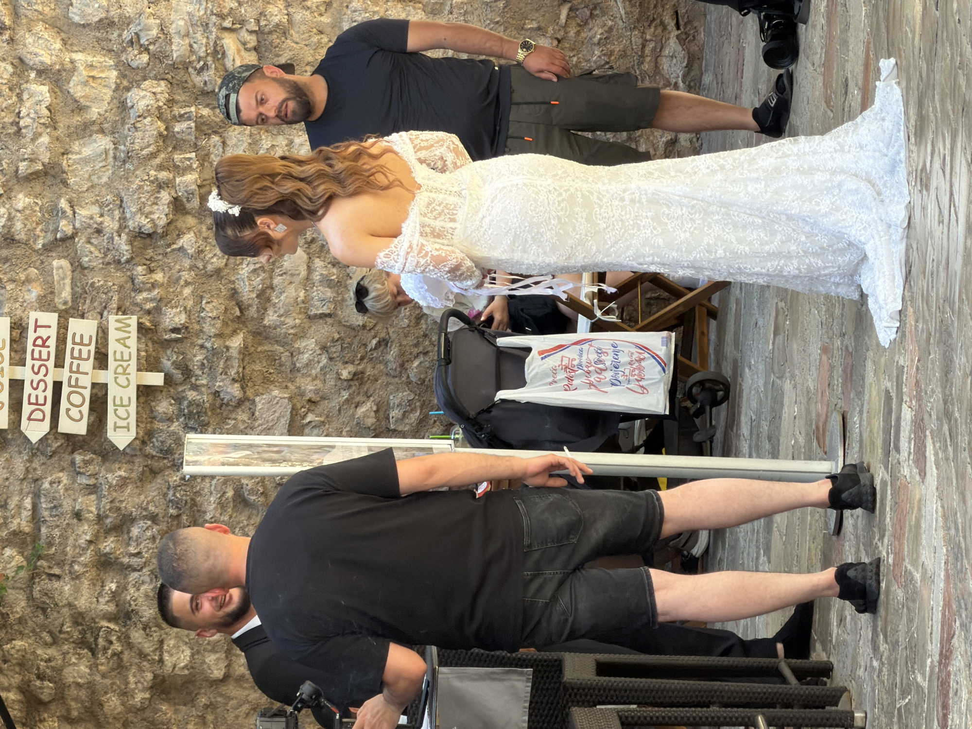 In Budva, Montenegro, a wedding party is gathered outdoors in front of a rough stone wall. A bride, wearing a white lace wedding dress with an off-the-shoulder neckline and a high ponytail adorned with a white flower, stands facing away from the camera, her back to the viewer. To her right, a man wearing a black t-shirt and olive-green cargo shorts, a camouflage baseball cap, and a gold watch, stands looking forward. To the bride's left, a man in a black t-shirt and black denim shorts, is partially visible, also facing away from the camera. Behind the bride, a woman with blonde hair wearing sunglasses is visible sitting. In the background, a white bag with French text and graphics is attached to a baby stroller. A directional signpost is mounted on the stone wall, with signs indicating "JUICE," "DESSERT," "COFFEE," and "ICE CREAM." The ground is paved with cobblestones.