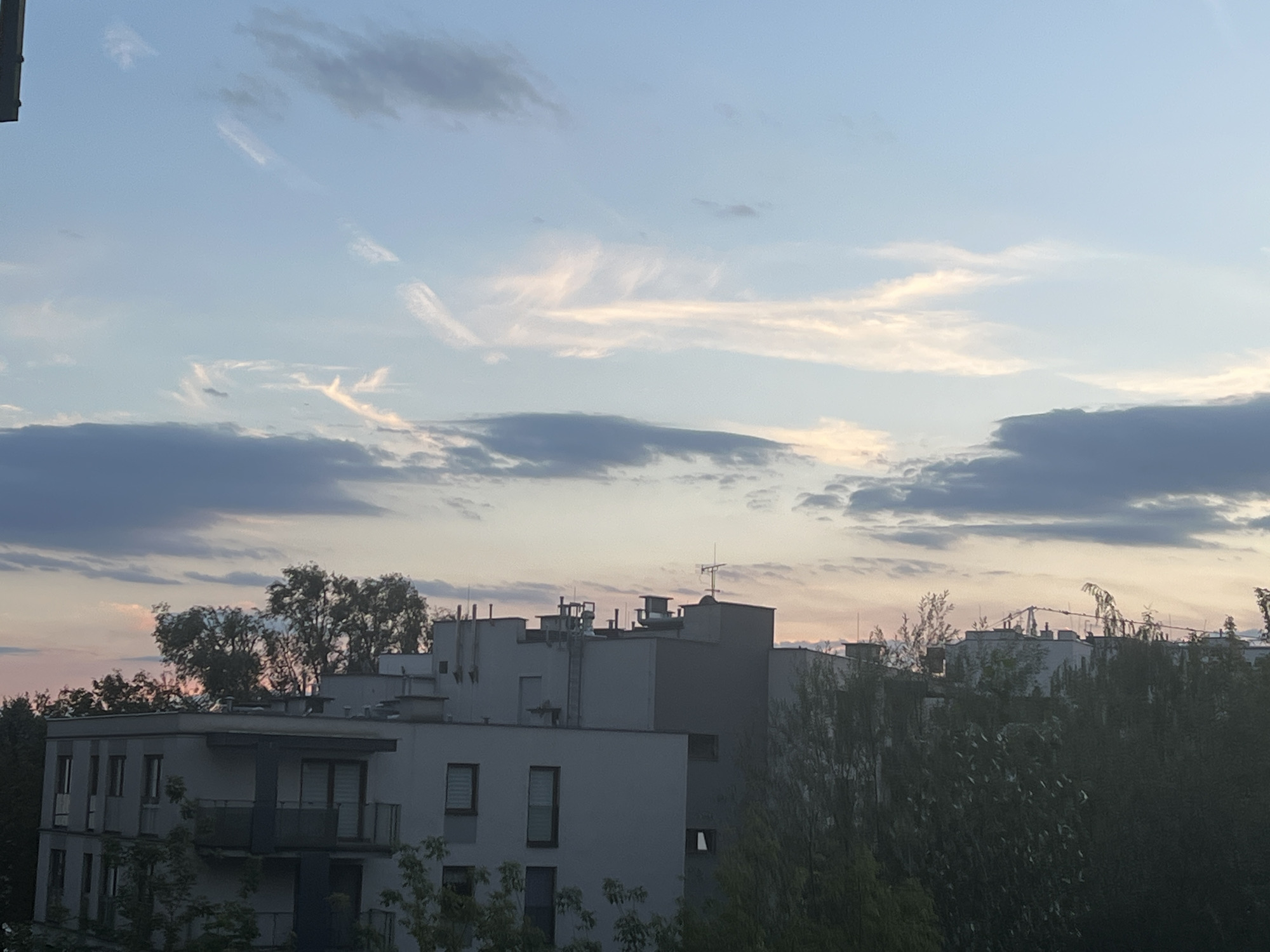 This is a photograph of an urban skyline at dusk, likely in Warsaw, Poland, given the architectural style of the buildings and the natural light conditions. The image captures a view looking towards the east, with the sky transitioning from a soft blue to shades of pink and orange near the horizon. Wispy, cirrus clouds are visible in the upper portion of the sky, interspersed with darker, more solid cloud formations lower down.
In the foreground and mid-ground, modern residential buildings are prominent. These structures are characterized by their flat roofs, multiple stories, and balconies. Some buildings feature large windows, some with visible blinds. Various rooftop elements, such as ventilation systems, pipes, and antennas, are apparent. Dense green foliage, consisting of trees and shrubs, partially obscures the lower sections of the buildings and extends across the scene, creating a layered effect. The lighting suggests it is either sunrise or sunset, with the light casting a gentle glow on the clouds and buildings. There are no people or vehicles visible in the frame. The overall atmosphere is calm and serene, characteristic of the evening or early morning.