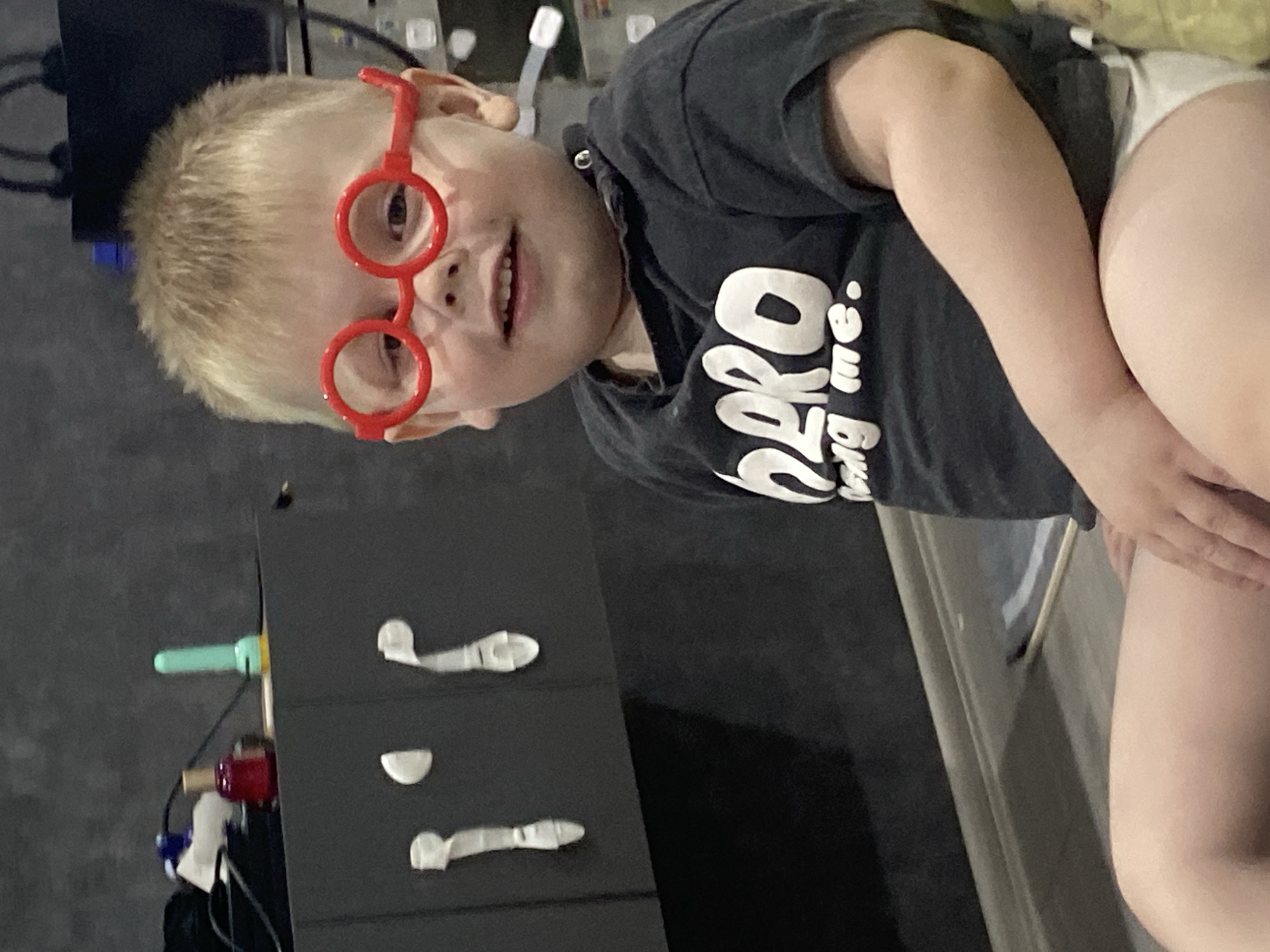 A young child with blond hair and wearing red, round-framed glasses is looking at the camera and smiling. They are wearing a dark grey t-shirt with white lettering and a white diaper. The child is positioned against a dark grey dresser or cabinet with silver-colored drawer pulls. Behind the dresser is a light-colored wall, and some cables and other items are on top of the dresser. The image provides no visual information to confirm the location as Chaikovsky, Russia.